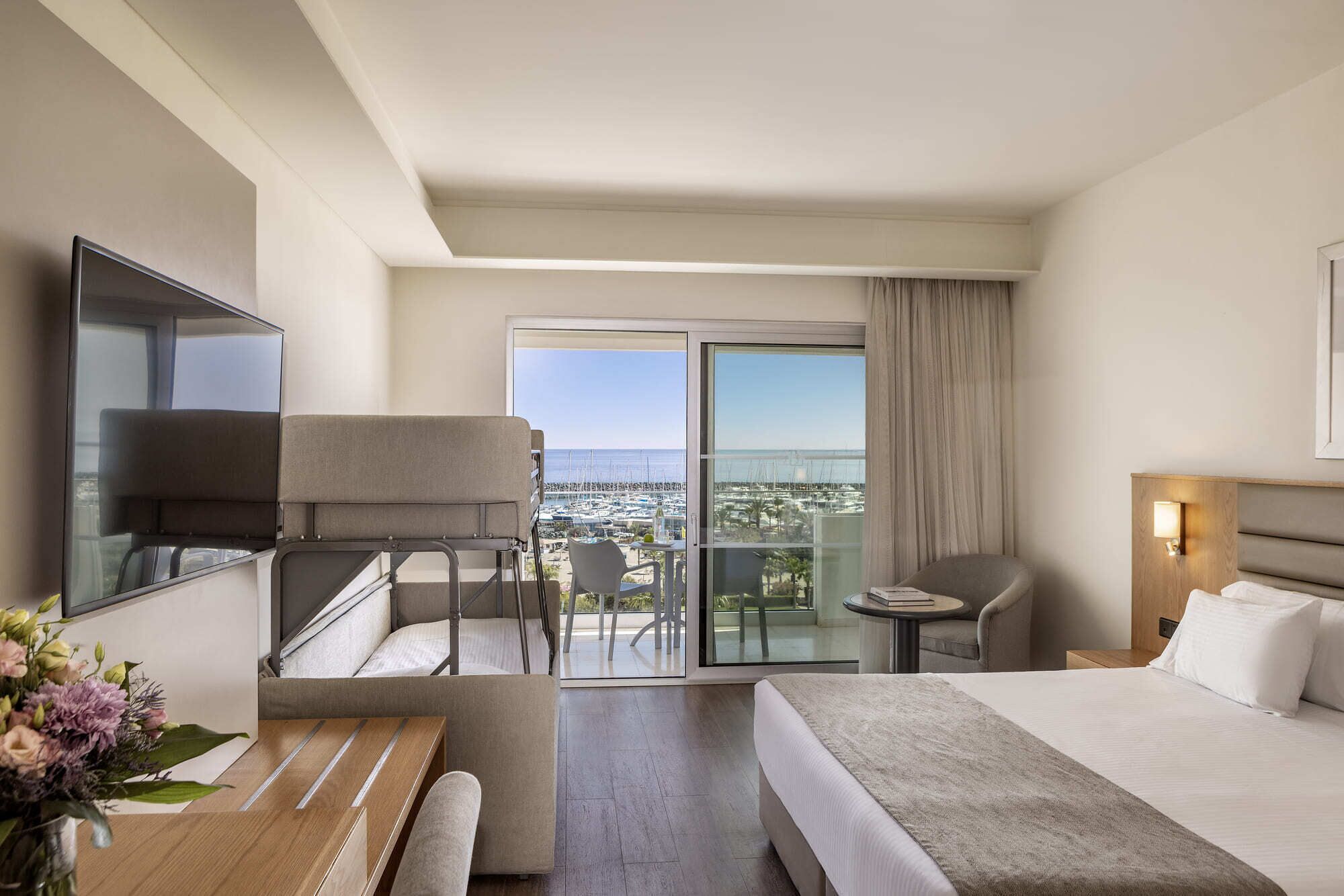 Habitación de hotel con cama, zona de estar con mesa y sillas, balcón con vista al mar y televisor montado en la pared.