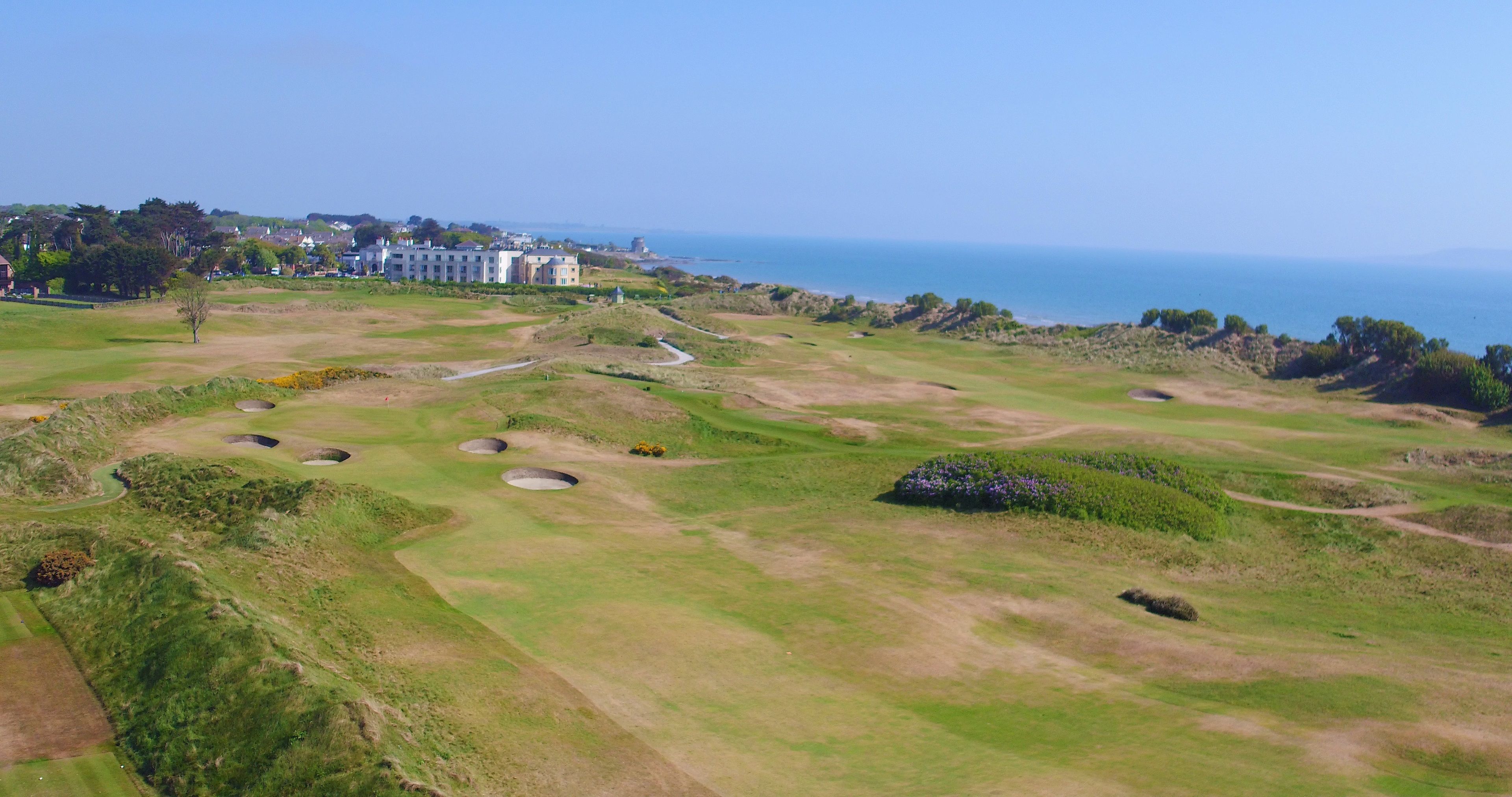 An aerial view of a golf course with bunkers, a clubhouse, and a view of the sea.