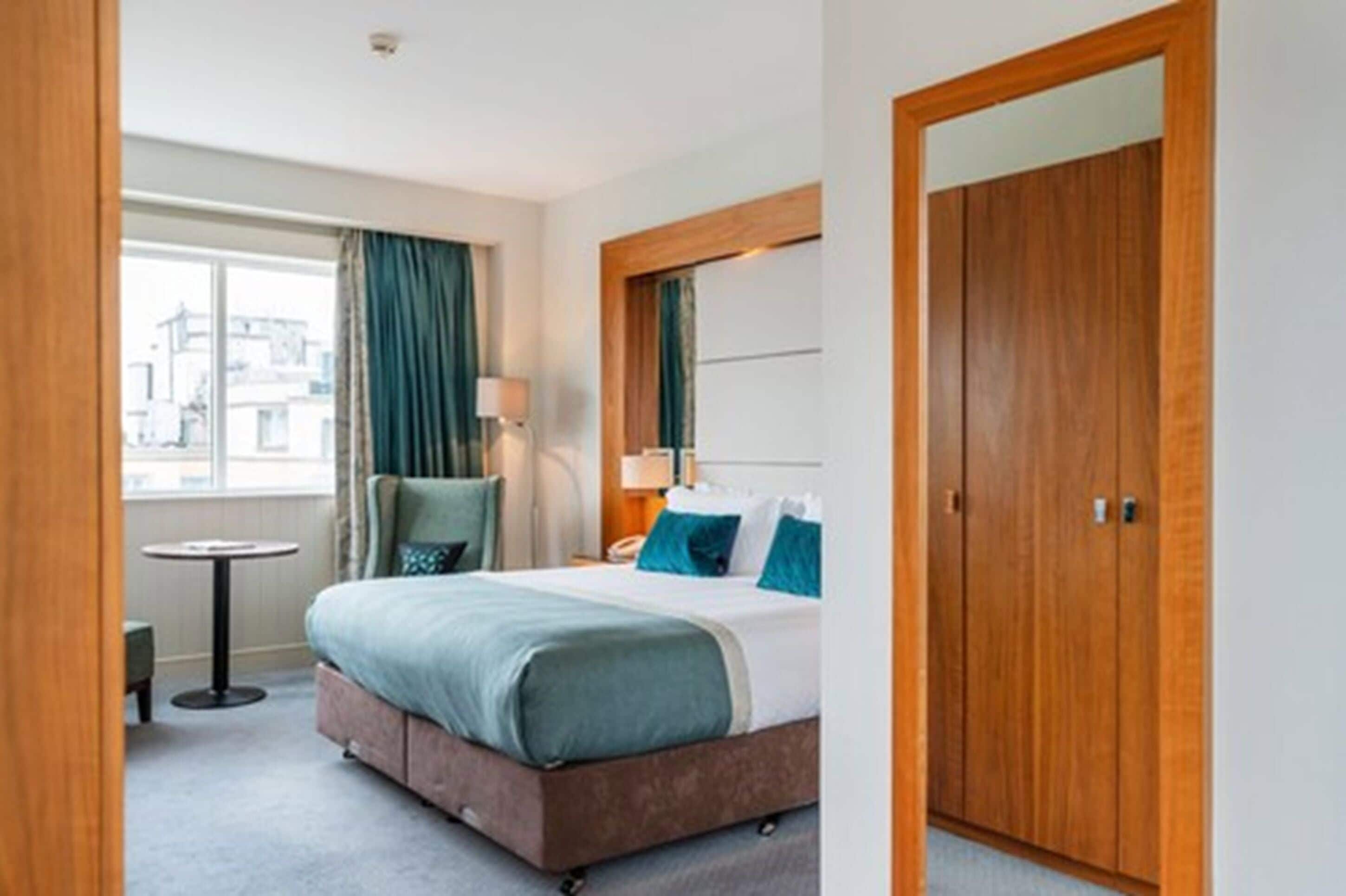 A hotel room with a bed, a round table, a chair, a lamp, and a wardrobe.