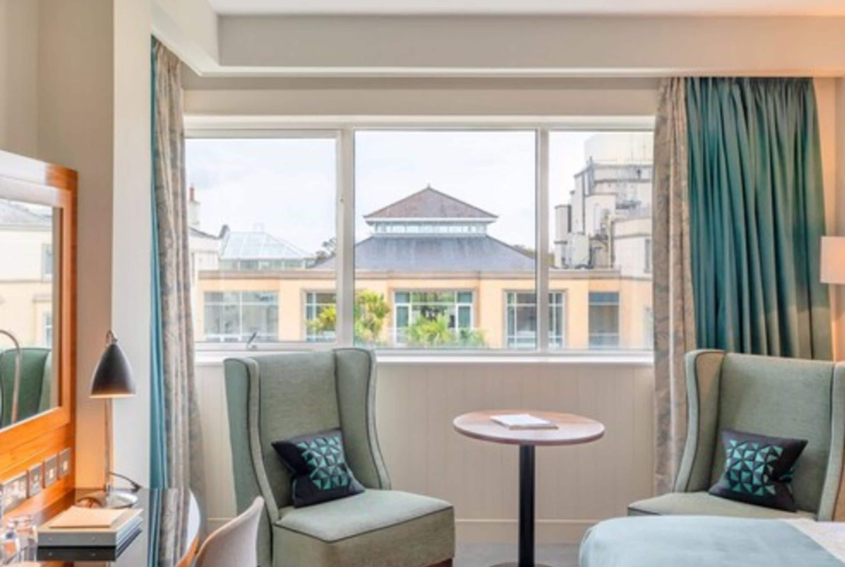 A hotel room with two green armchairs, a small round table, and a view of buildings through large windows.