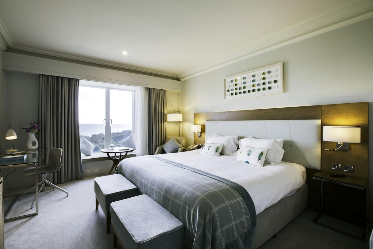 A hotel room with a large bed, a desk with a chair, a small table, and a bench. There are two bedside tables with lamps, a wall-mounted artwork, and a view of the outdoors through a window.