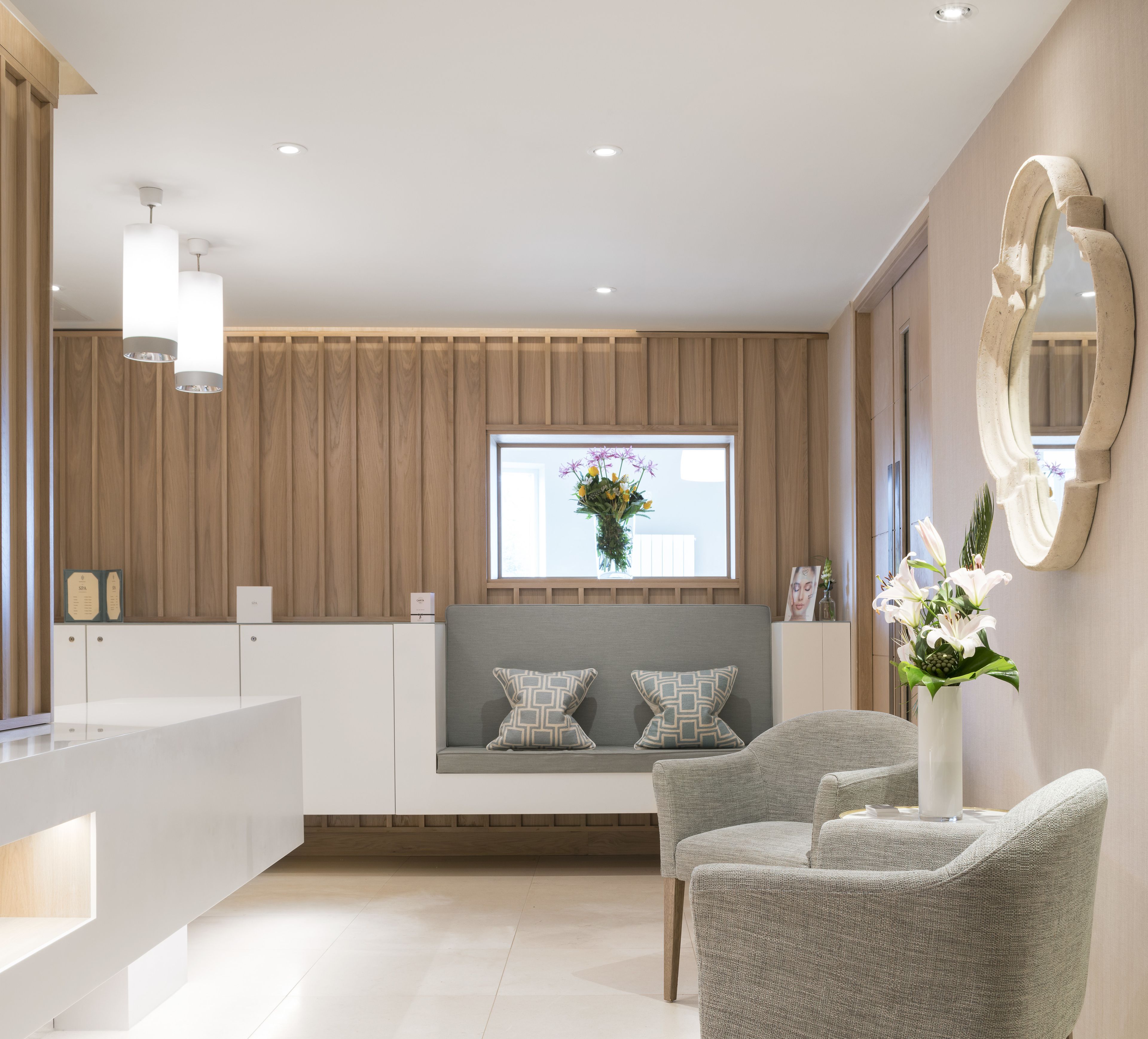 A modern hotel room with a wooden panel wall, a built-in bench with cushions, a large mirror, and a seating area with grey upholstered chairs.