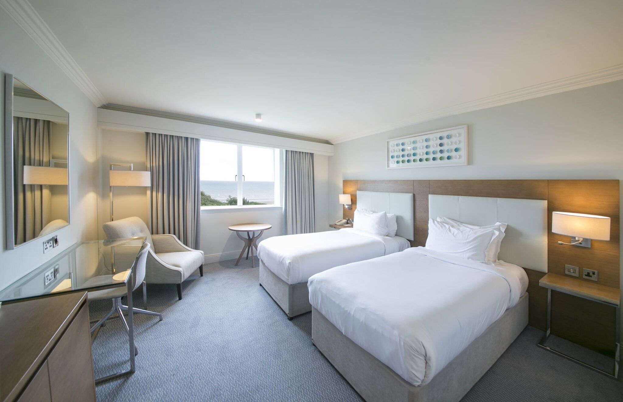 A hotel room with two beds, a desk, a chair, a small table, and a view of the sea.