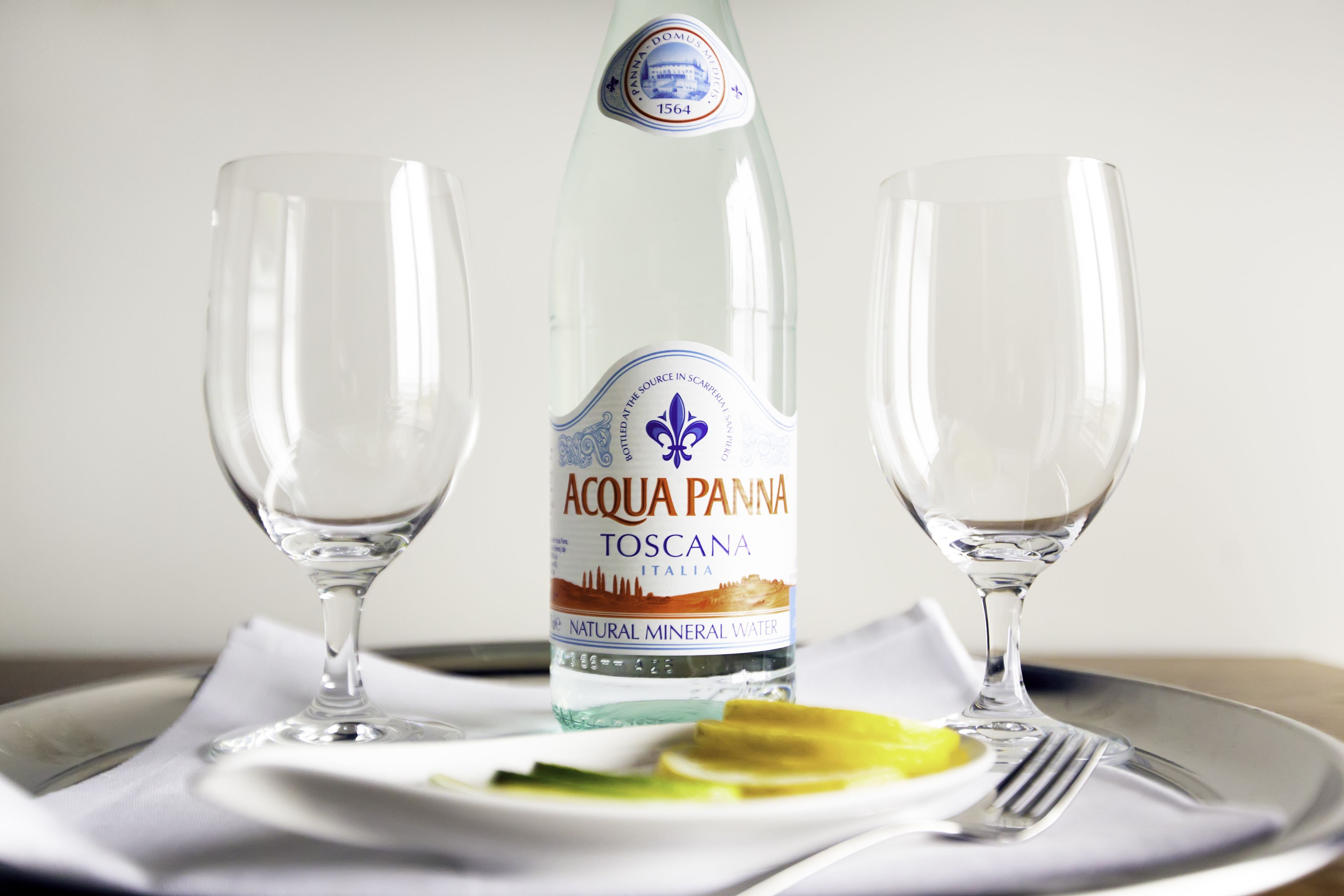 A bottle of Acqua Panna Toscana water, two empty wine glasses, a plate with a slice of lemon, and a fork on a tray.
