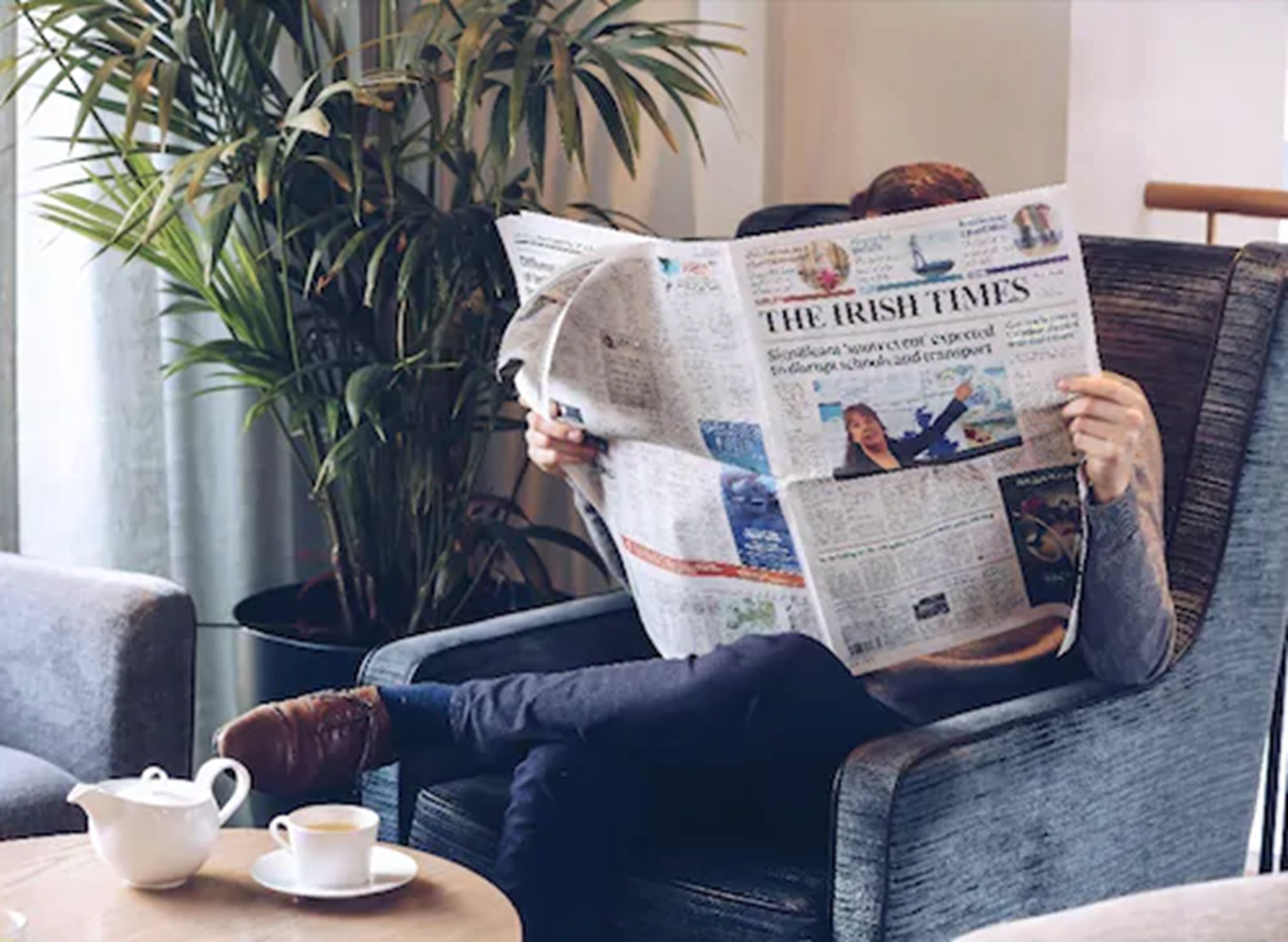 A person reading a newspaper in a room with a plant and a table with a teapot and cup.