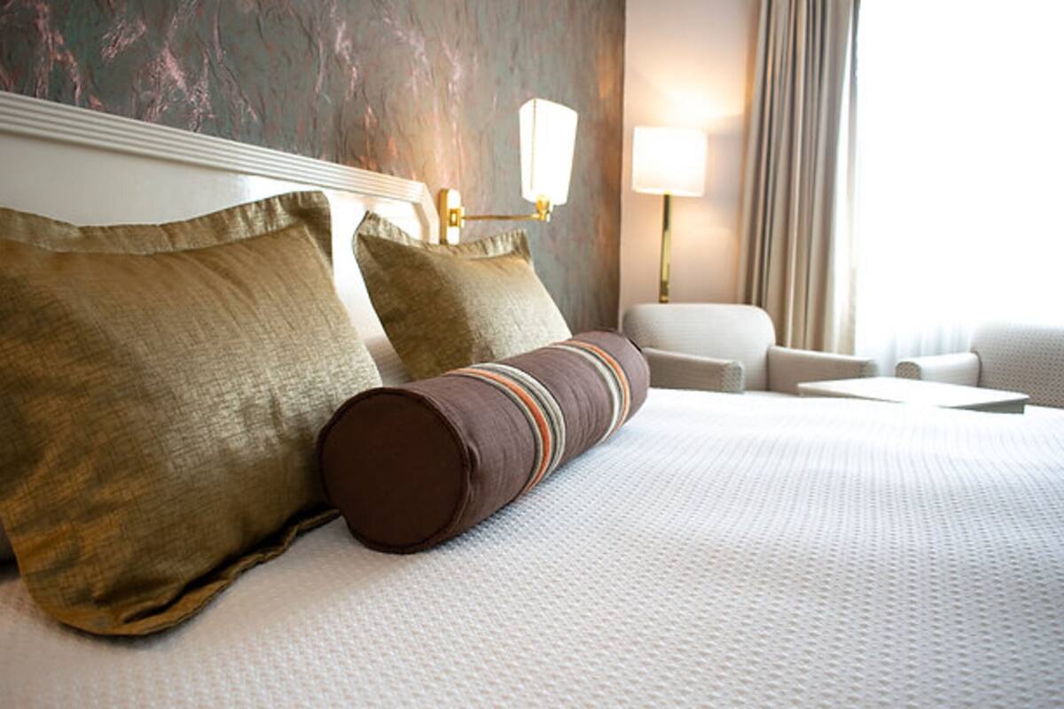 Una cama bien hecha con un edredón blanco y dos almohadas grandes, una almohada cilíndrica marrón y una mesita de noche con una lámpara.
