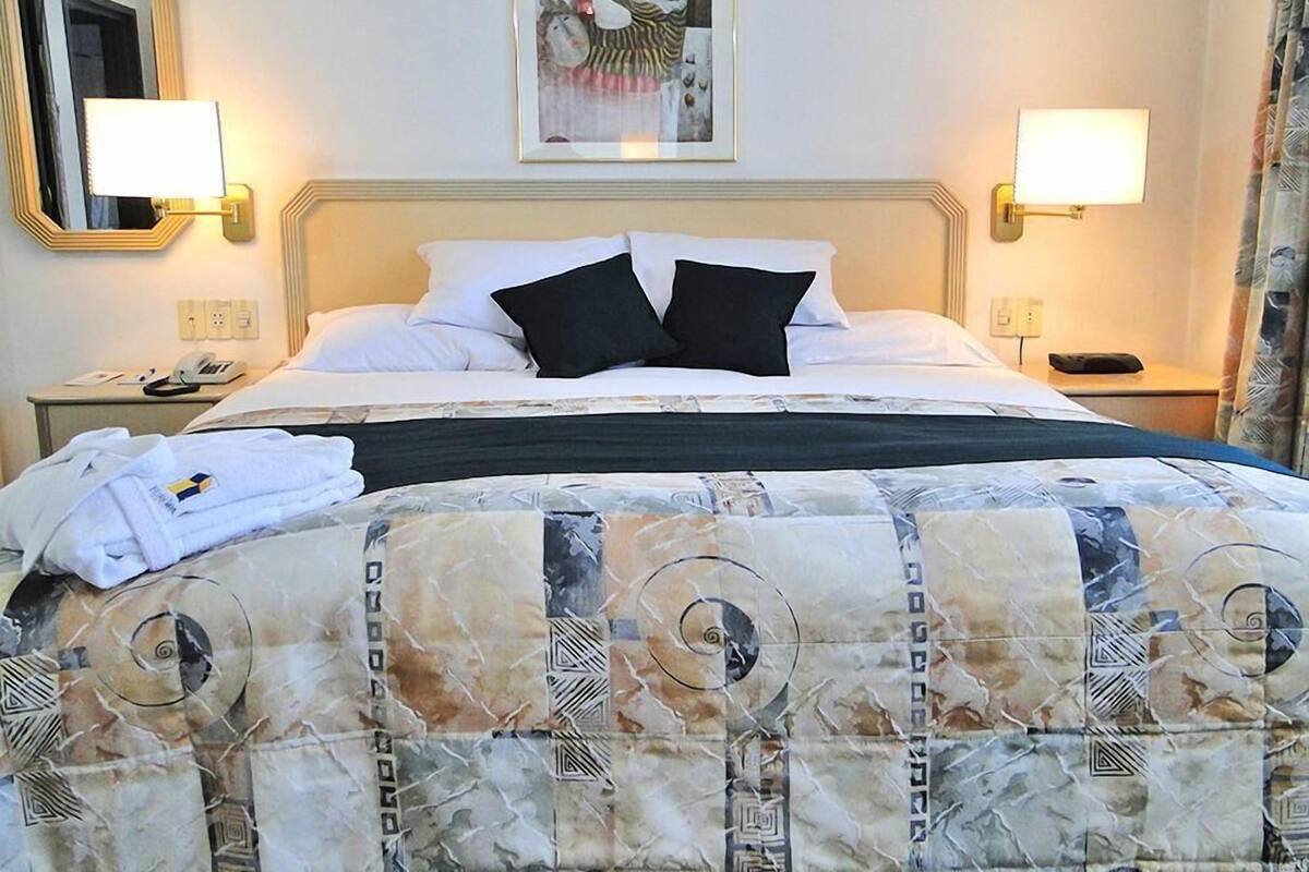 Habitación de hotel con cama, dos lámparas de noche, un cuadro enmarcado sobre la cama y un teléfono en la mesita.