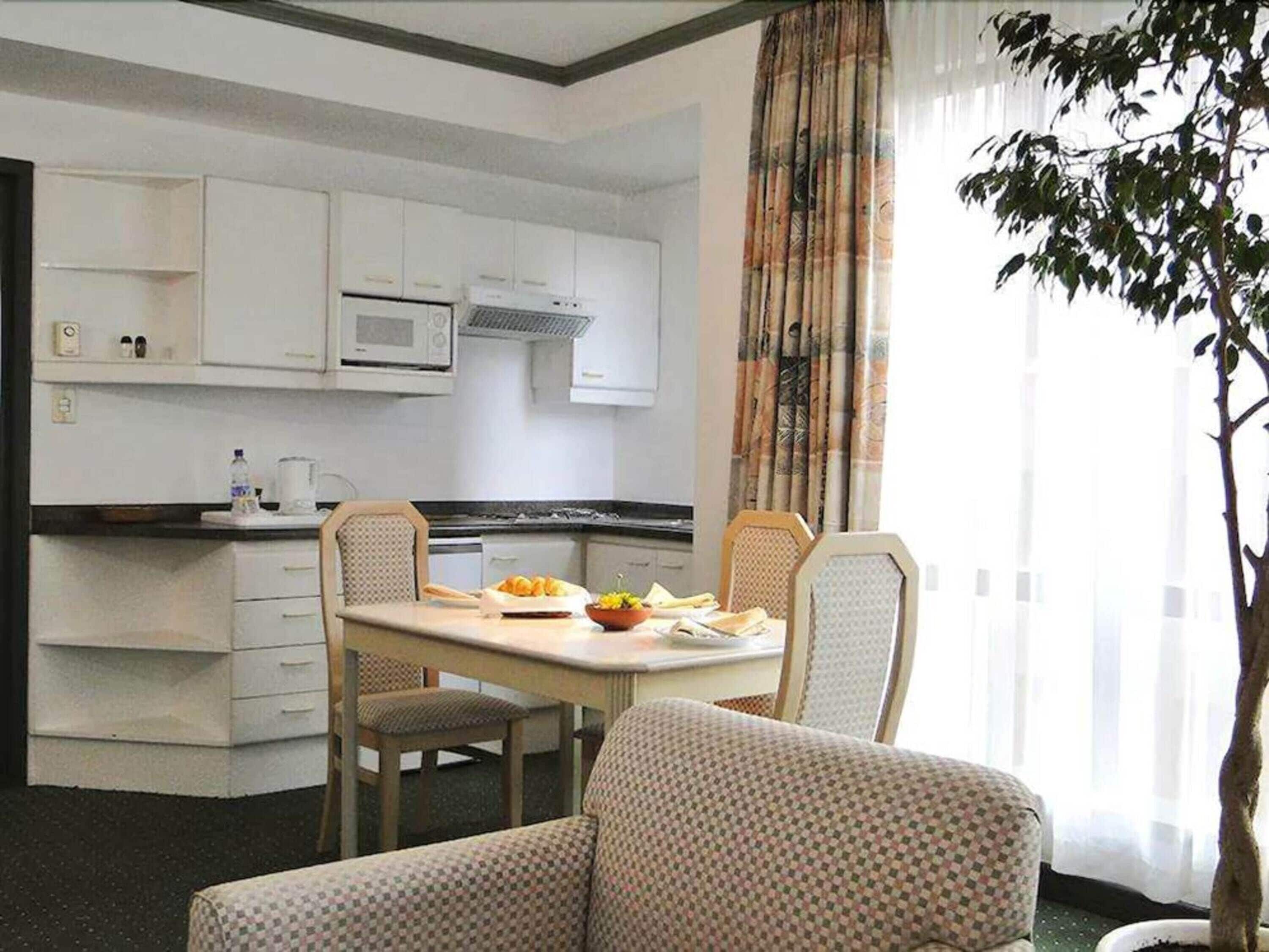 A compact hotel room with a kitchenette, a dining area with a table and chairs, and a large window with curtains.