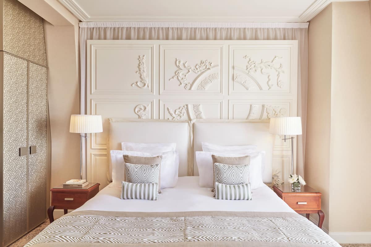 Quarto de hotel com uma cama grande, duas mesas de cabeceira, cabeceira com painéis decorativos e mesa de apoio com abajur.