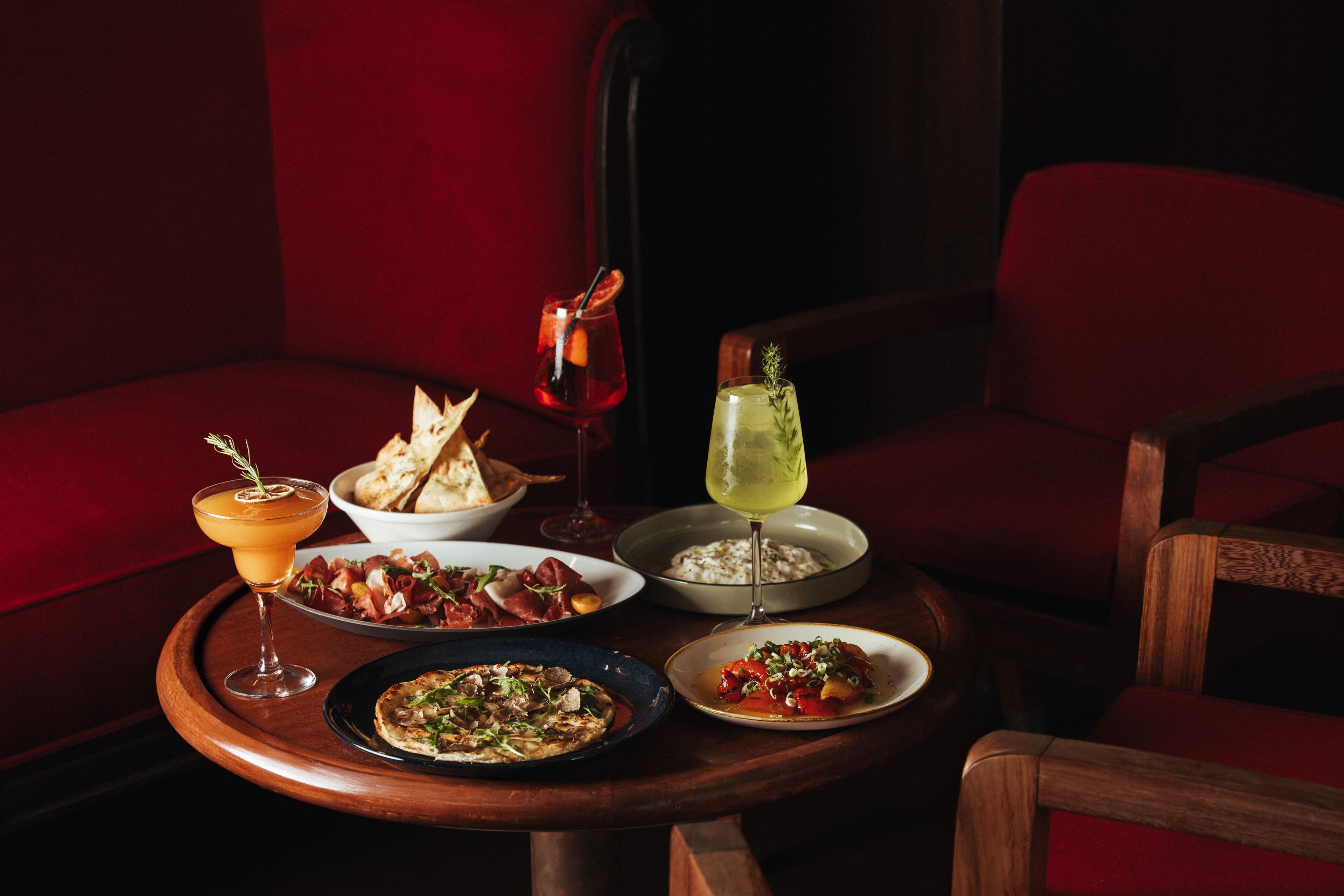 Una mesa de comedor con diversos platos y bebidas, en un ambiente tenuemente iluminado y con asientos tapizados en rojo.