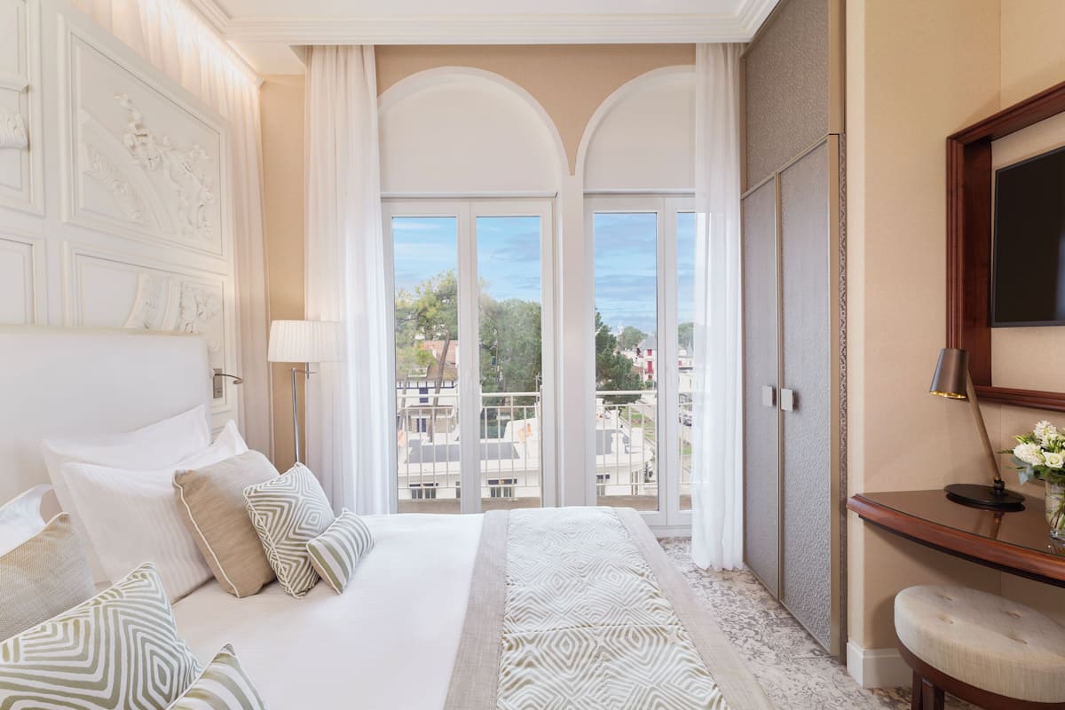 Quarto de hotel com uma cama grande, vista através de uma varanda e uma escrivaninha de madeira com abajur.