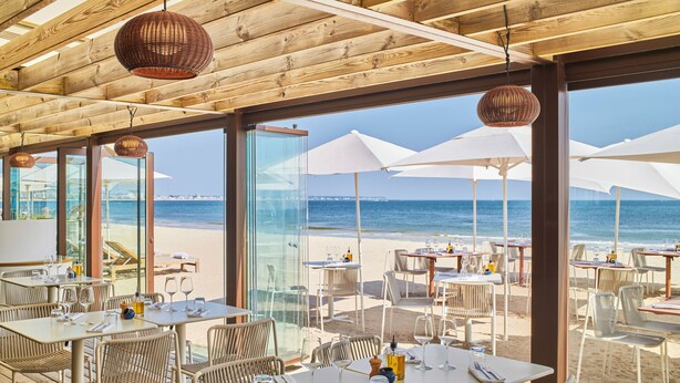 Un área de comedor al aire libre con mesas y sillas, grandes sombrillas y vistas a la playa y al océano.