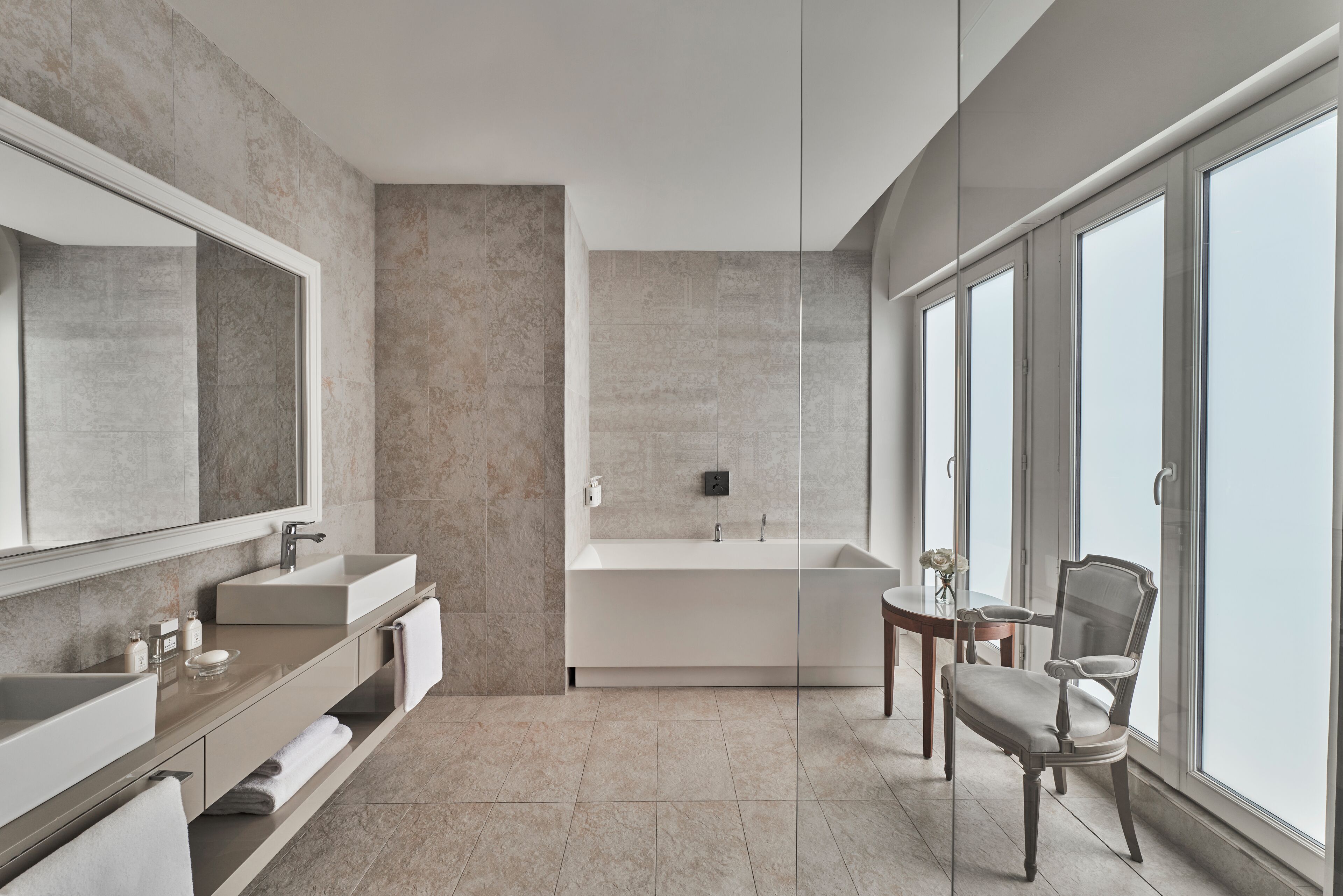 A modern bathroom with a large mirror, two sinks, a bathtub, and a chair by a large window.