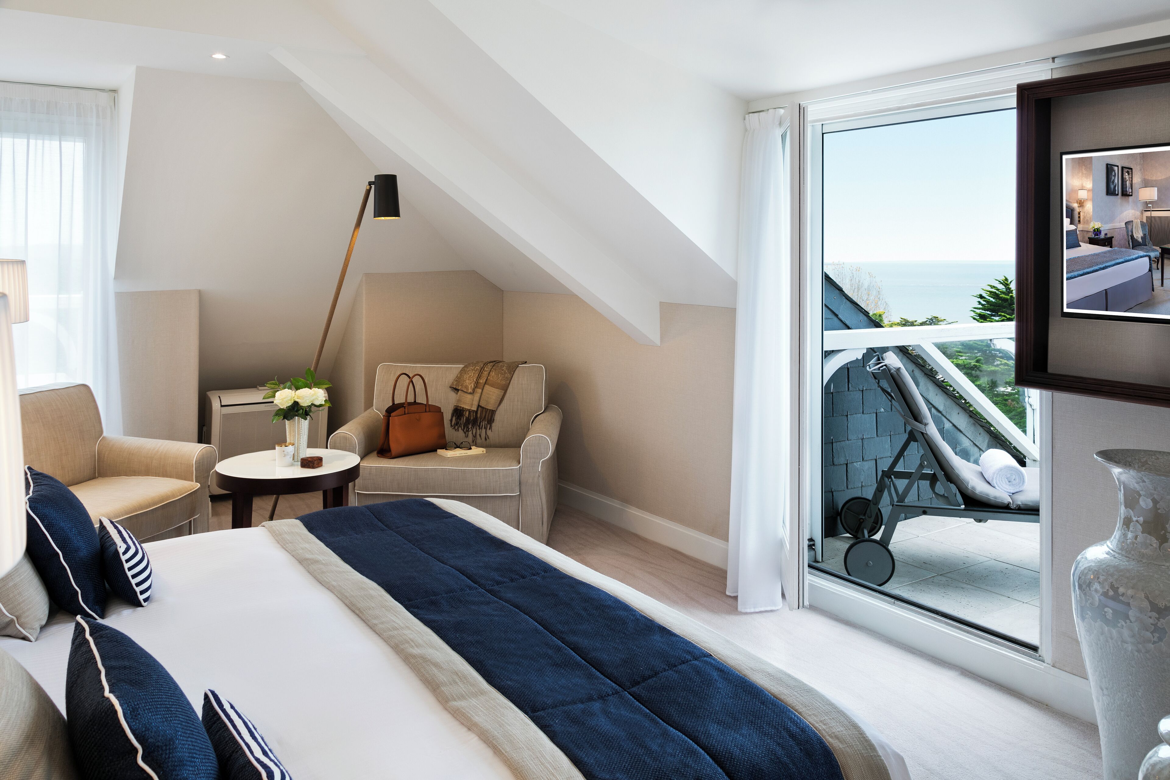 A modern hotel room with a large bed, a seating area with a table, and a balcony with a view of the sea.