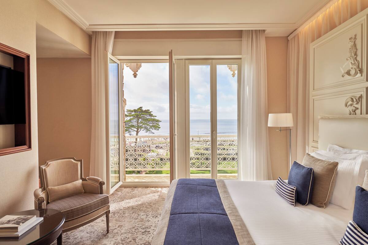 Quarto de hotel com uma cama grande, uma cadeira, uma mesa e vista para o exterior através de uma porta de correr.
