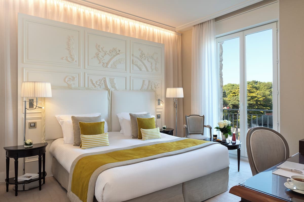 Quarto de hotel com uma cama grande, duas mesas de cabeceira com luminárias, uma cadeira, uma pequena mesa com um vaso e uma varanda com vista para árvores.