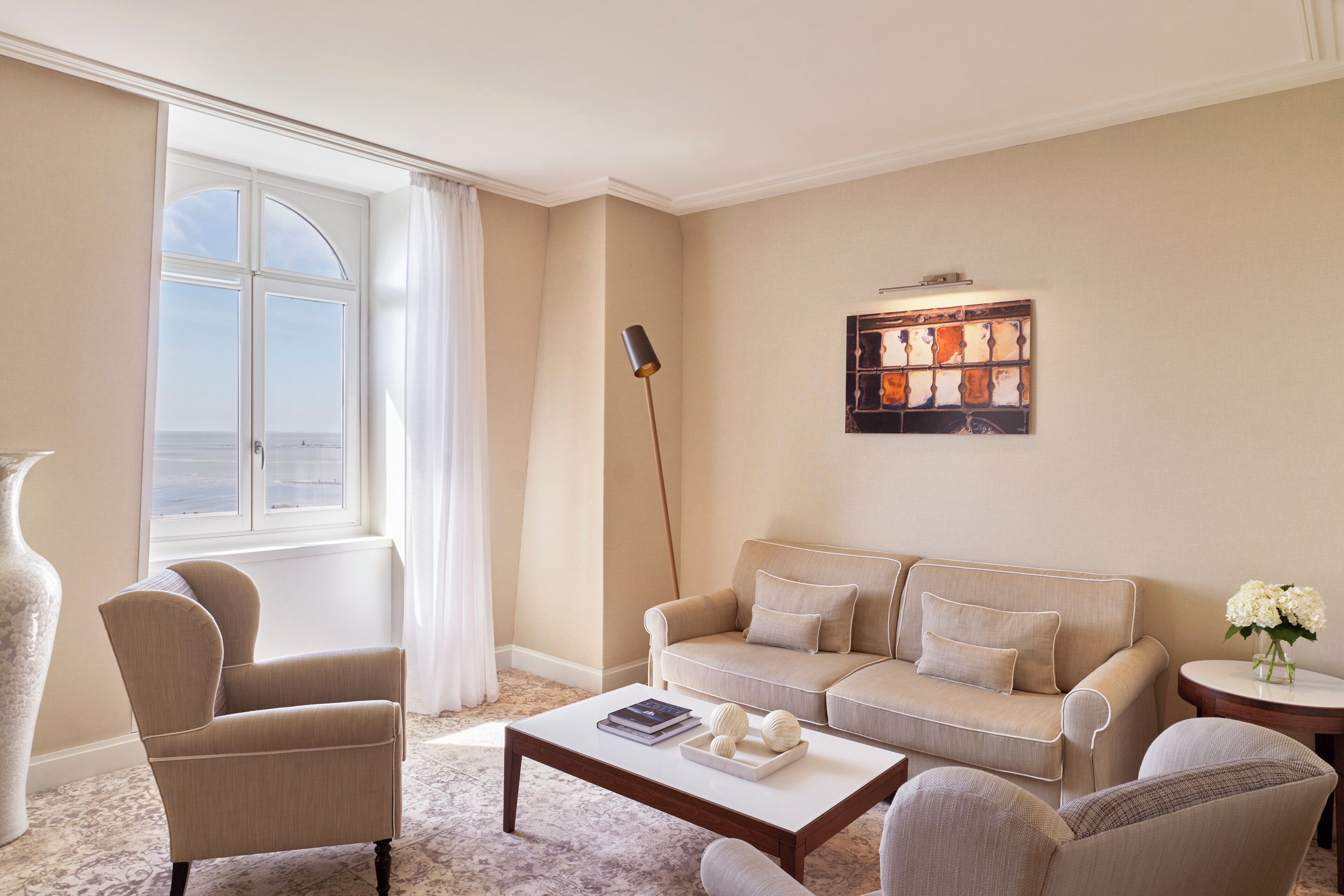 En stue med en beige sofa, et sofabord og en lænestol. Der er et vindue med gardiner, en gulvlampe og et billede på væggen.