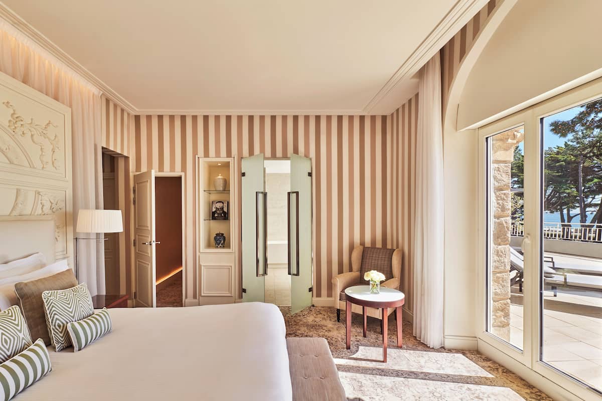 Quarto de hotel com uma cama grande, uma mesa redonda pequena, uma cadeira, um guarda-roupa e vista para o exterior.