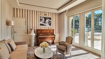Una sala de estar con sofá, sillón, piano y un ventanal con vistas a árboles.