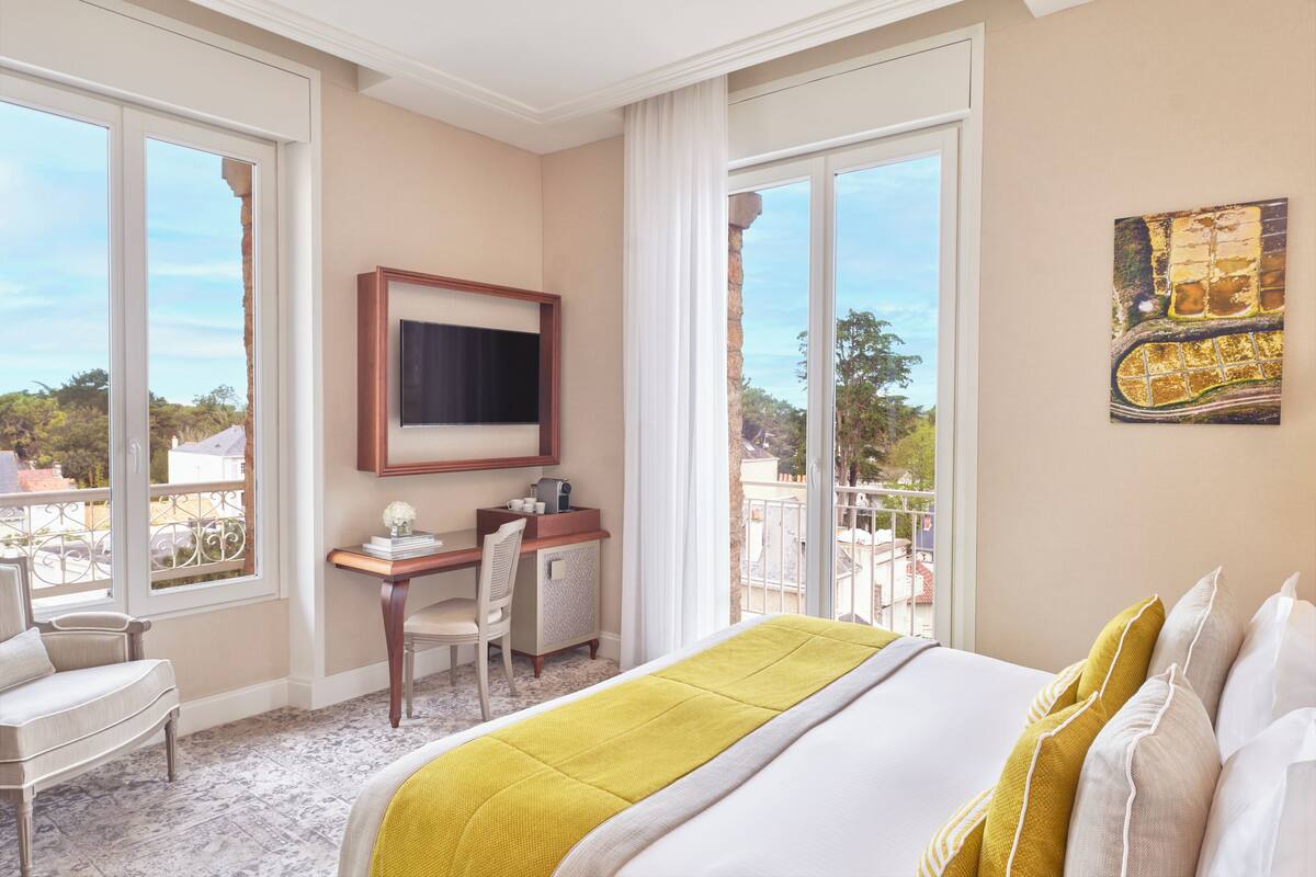 Quarto de hotel com cama, escrivaninha, cadeira, televisão e grandes janelas com vista para o exterior.