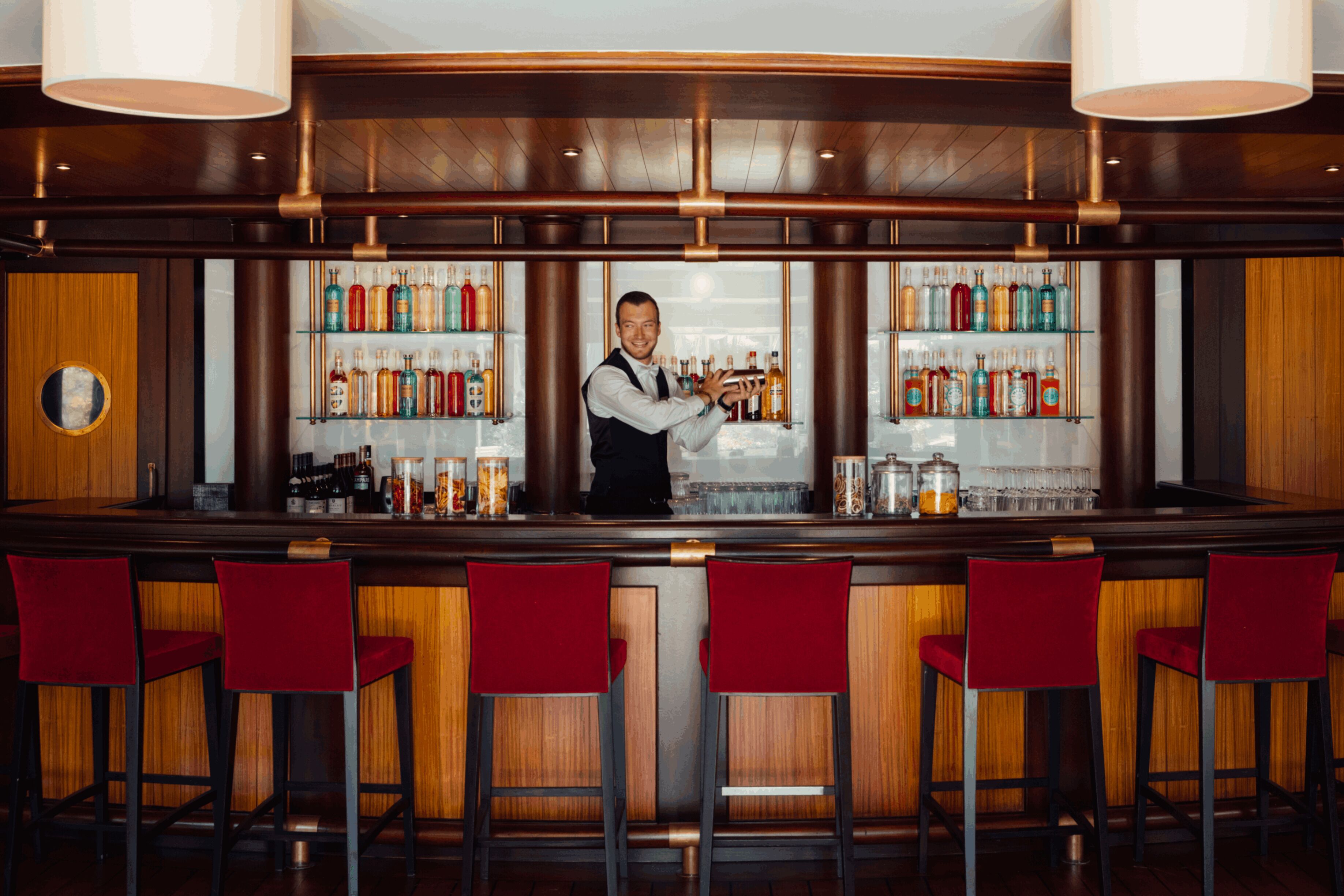 Zona de bar con un cantinero, taburetes rojos y estantes llenos de botellas de licor.