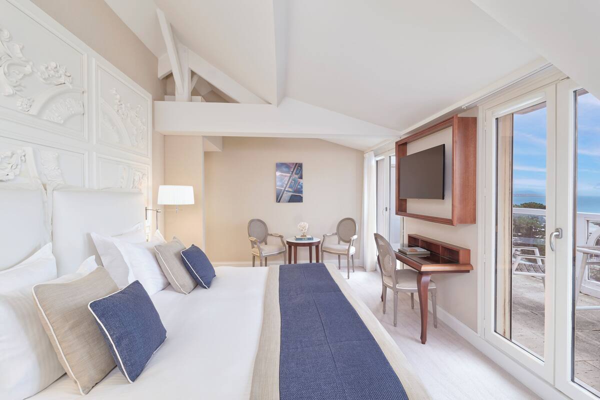 Quarto de hotel com cama, escrivaninha, televisão e varanda com área de estar.