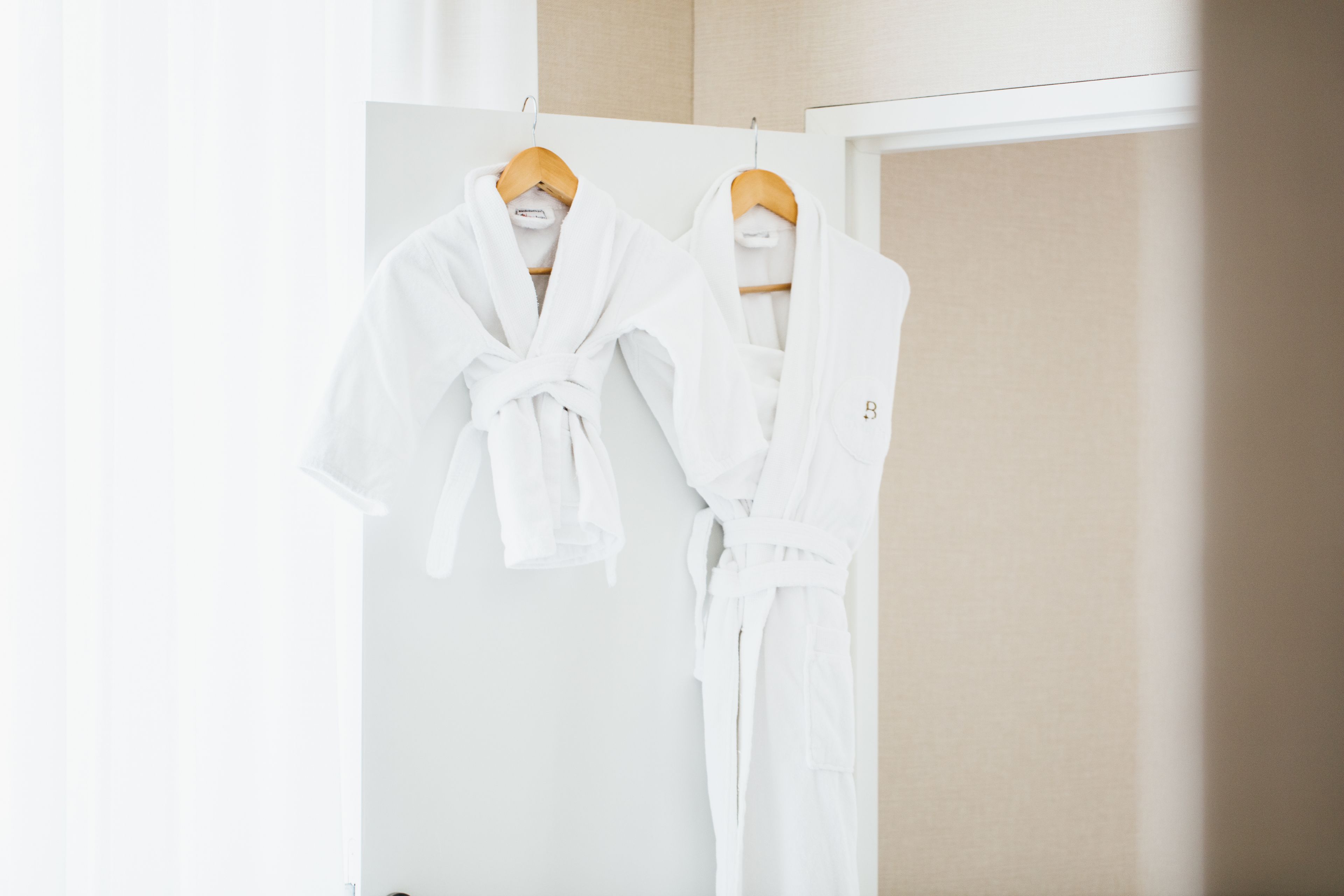 Deux robes blanches sont suspendues à des cintres en bois dans une pièce dont le mur est de couleur beige et la porte, blanche.