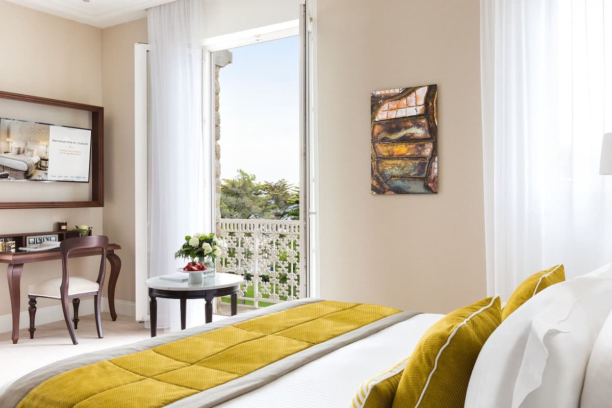 Quarto de hotel com uma cama, uma escrivaninha, uma cadeira, uma mesa pequena com um vaso de flores e um quadro na parede.