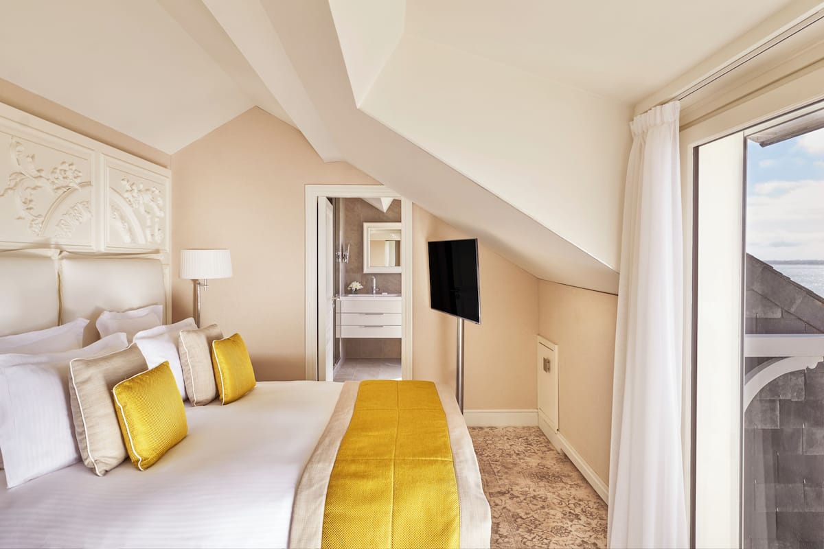 Quarto de hotel com uma cama grande, televisão, banheiro visível por uma porta aberta e varanda com vista.