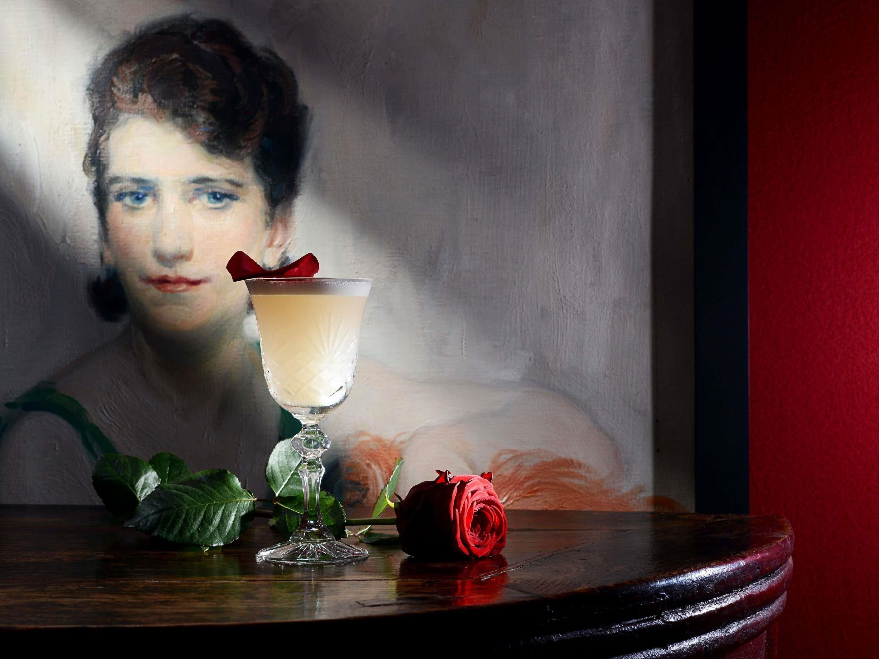 Une table avec un verre contenant une boisson de couleur claire, une rose rouge et des feuilles vertes.