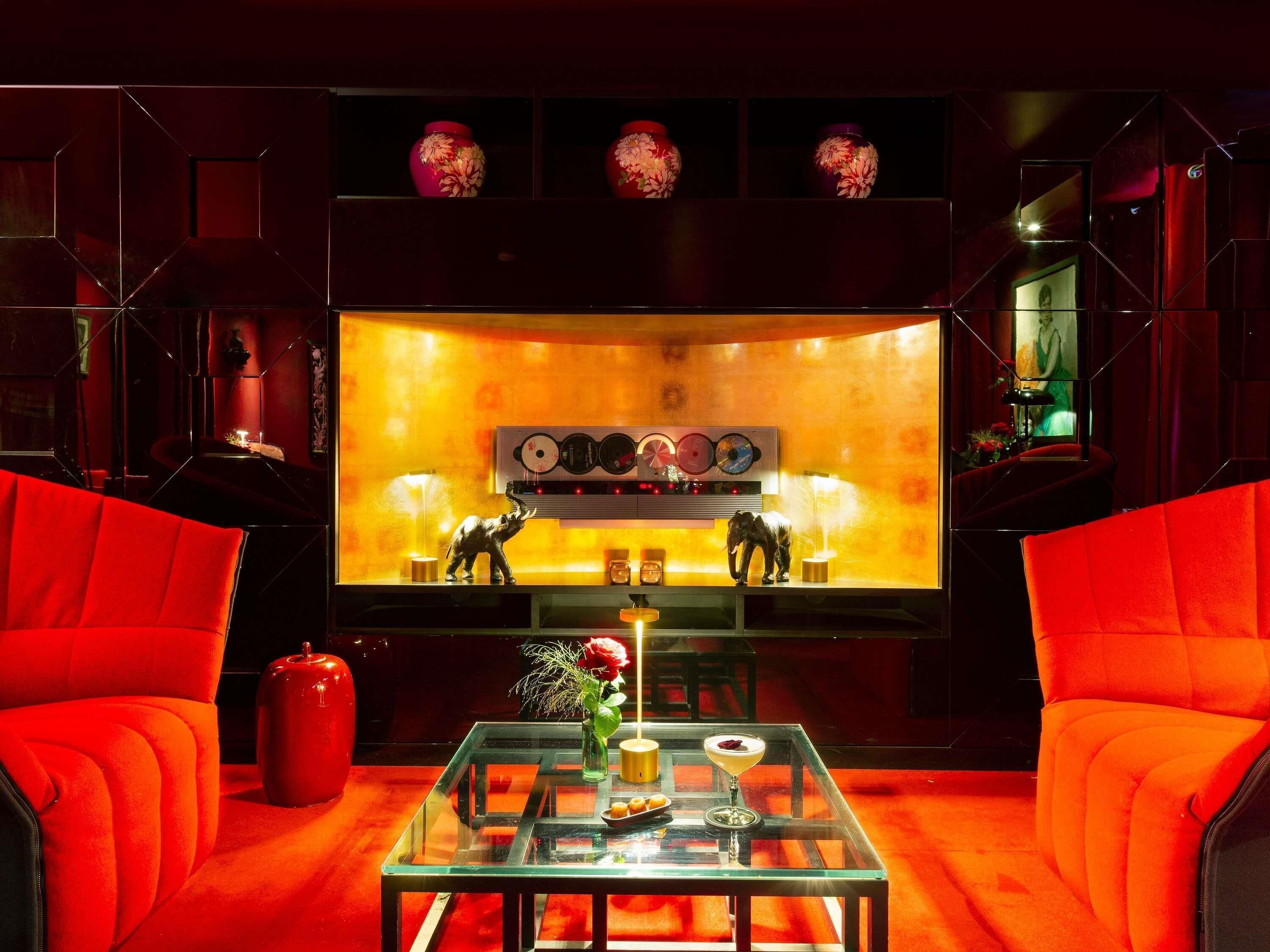 A room with red upholstered chairs, a glass coffee table with a vase and a drink, and a wall with decorative vases and a record player.