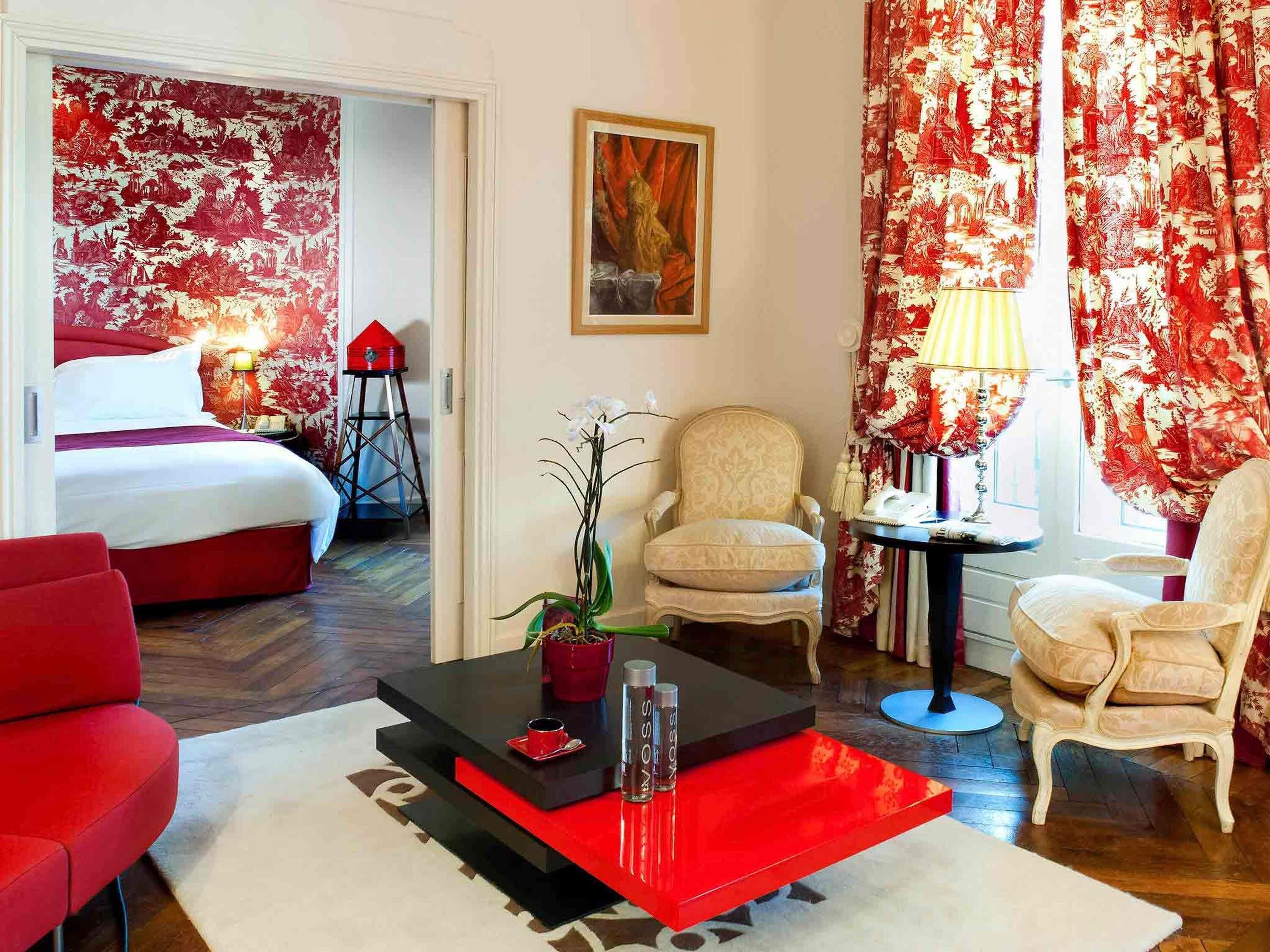 A hotel room with a bed, a red and black coffee table, a white armchair, a red armchair, and a small table with a lamp.