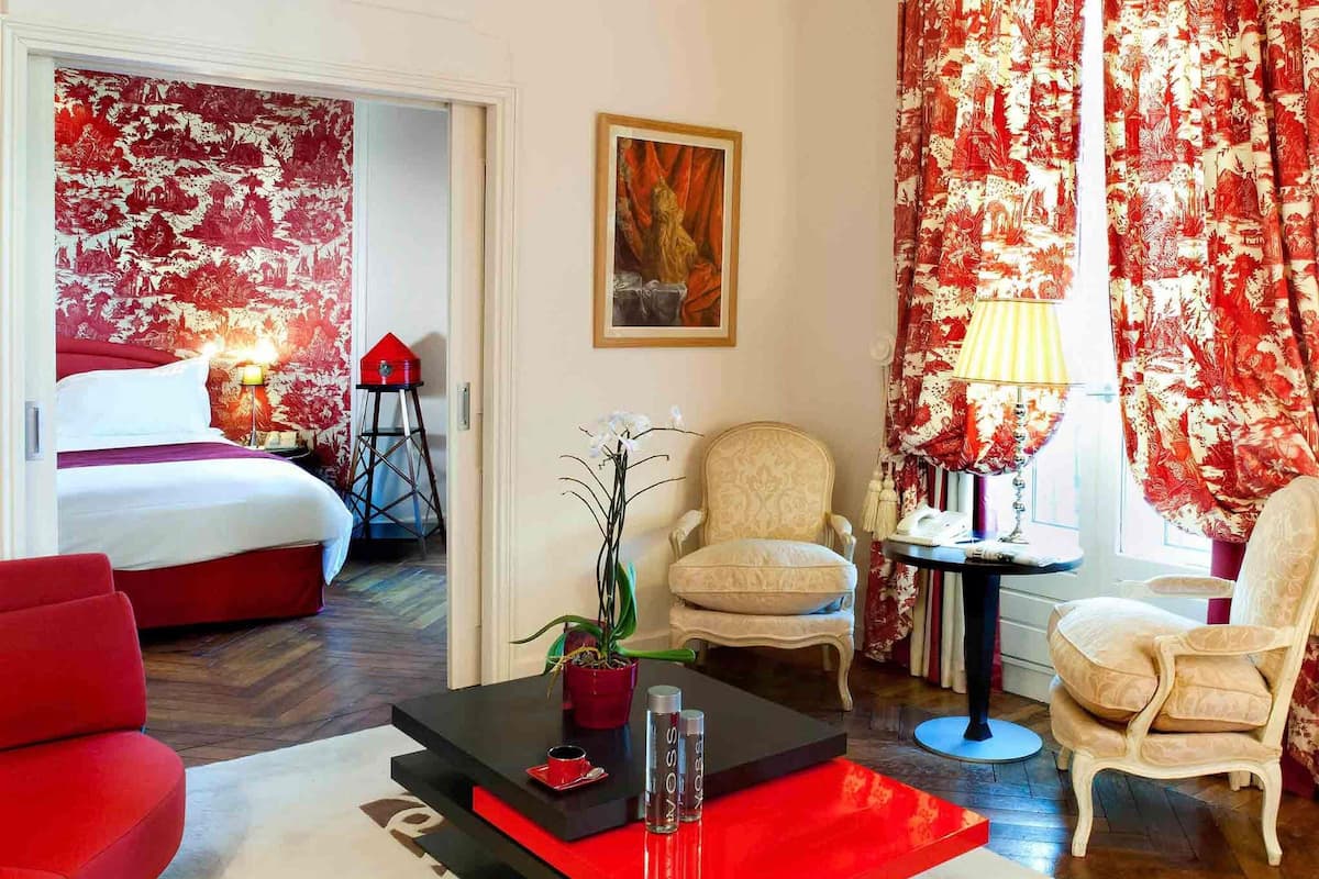 Une chambre d’hôtel avec un lit, une table basse rouge et noire, un fauteuil blanc, un fauteuil rouge et une petite table avec une lampe.