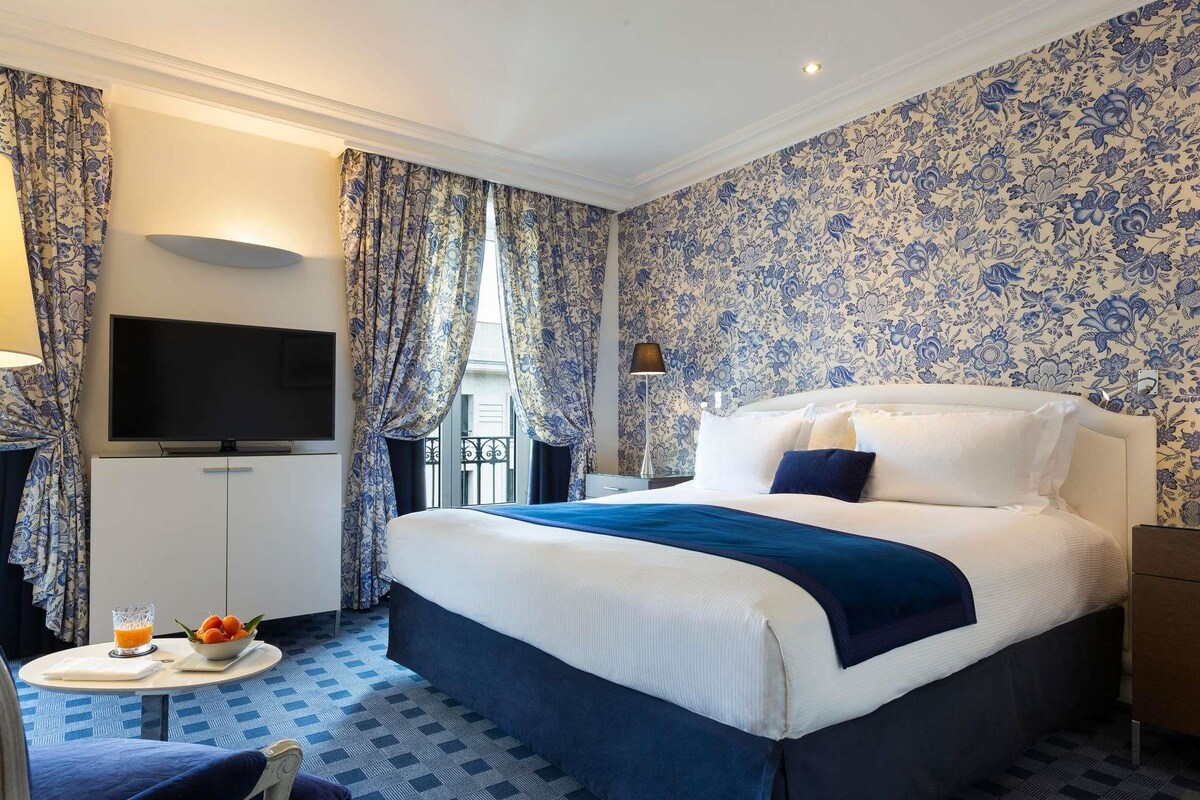 A hotel room with a bed, a TV, a desk with a lamp, and a chair.