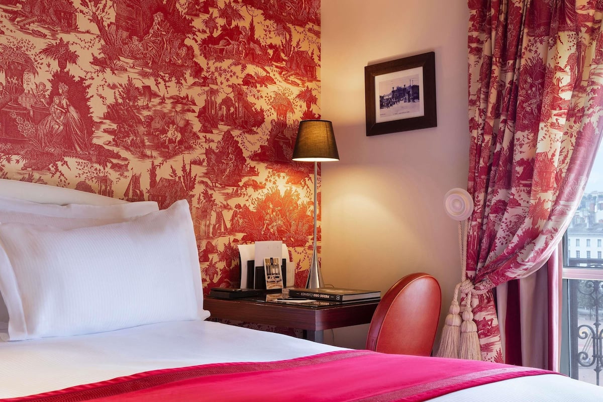 Une chambre d’hôtel avec un lit, un bureau, une lampe, une chaise et une fenêtre avec des rideaux.
