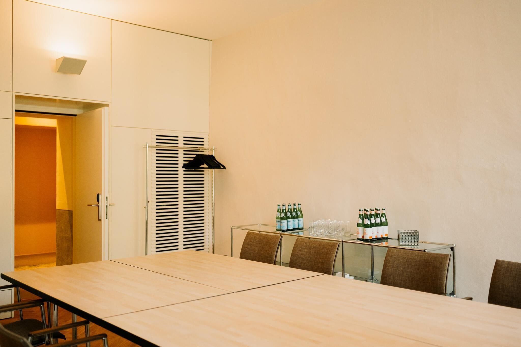 A conference room with a long table, chairs, a glass-top table, and a cabinet with bottles and glasses.
