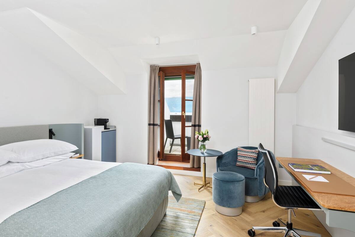 A modern hotel room with a bed, a desk, and a chair. There is a view of the outdoors through a door.