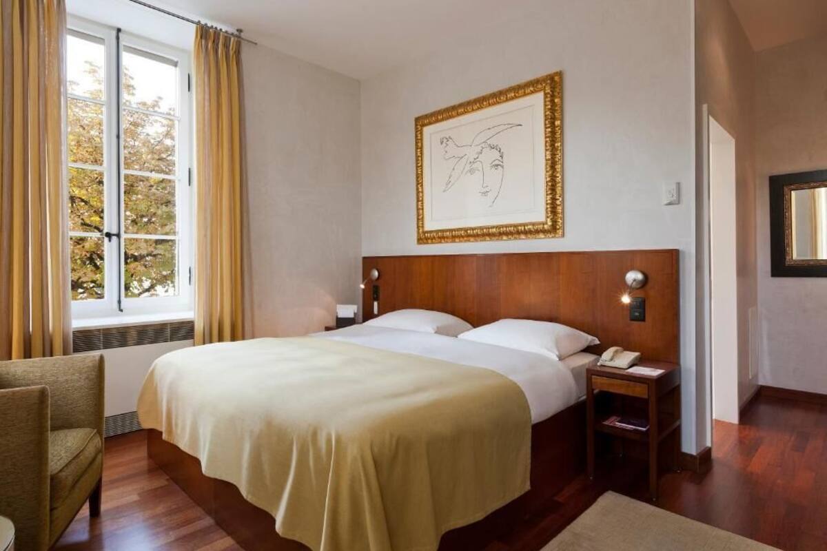 A hotel room with a large bed, a bedside table with a lamp, a framed artwork on the wall, and a window with curtains.