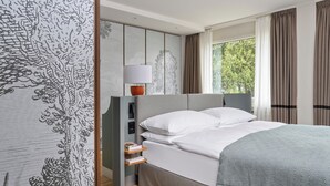 Une chambre d’hôtel moderne avec un grand lit, une table de chevet avec des livres, une lampe et une vue sur la verdure.
