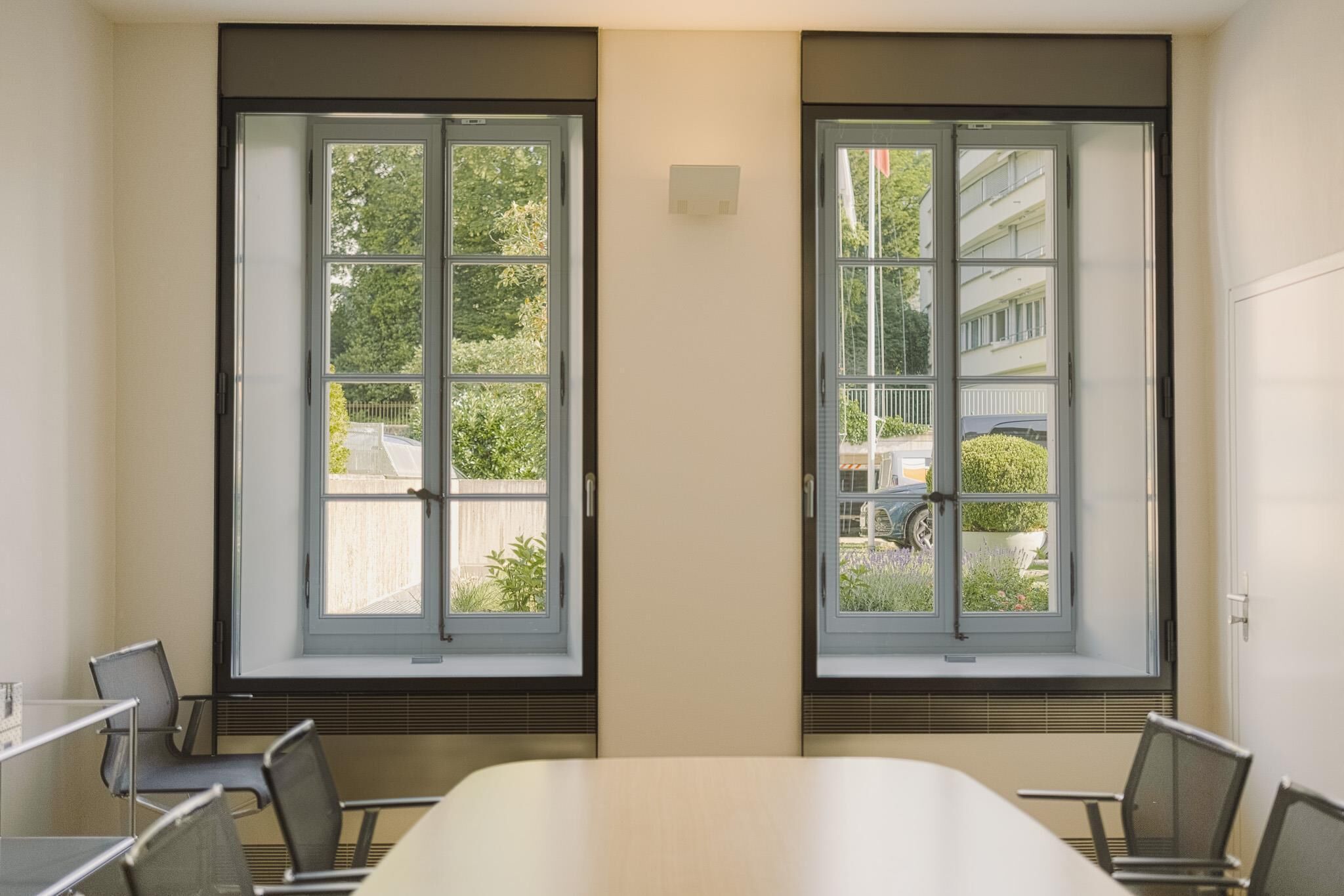 A conference room with a long table and four chairs, large windows with a view of greenery and buildings, and a whiteboard on the wall.