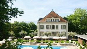 Un grand bâtiment doté d’une piscine, entouré d’une végétation luxuriante et comprenant un espace de détente extérieur.