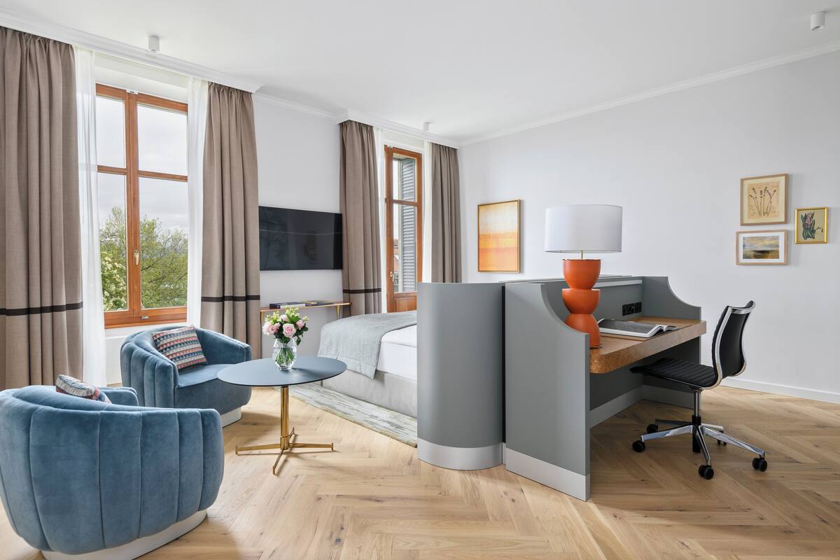 A modern hotel room with a bed, a desk, a blue armchair, a small table, and a TV.