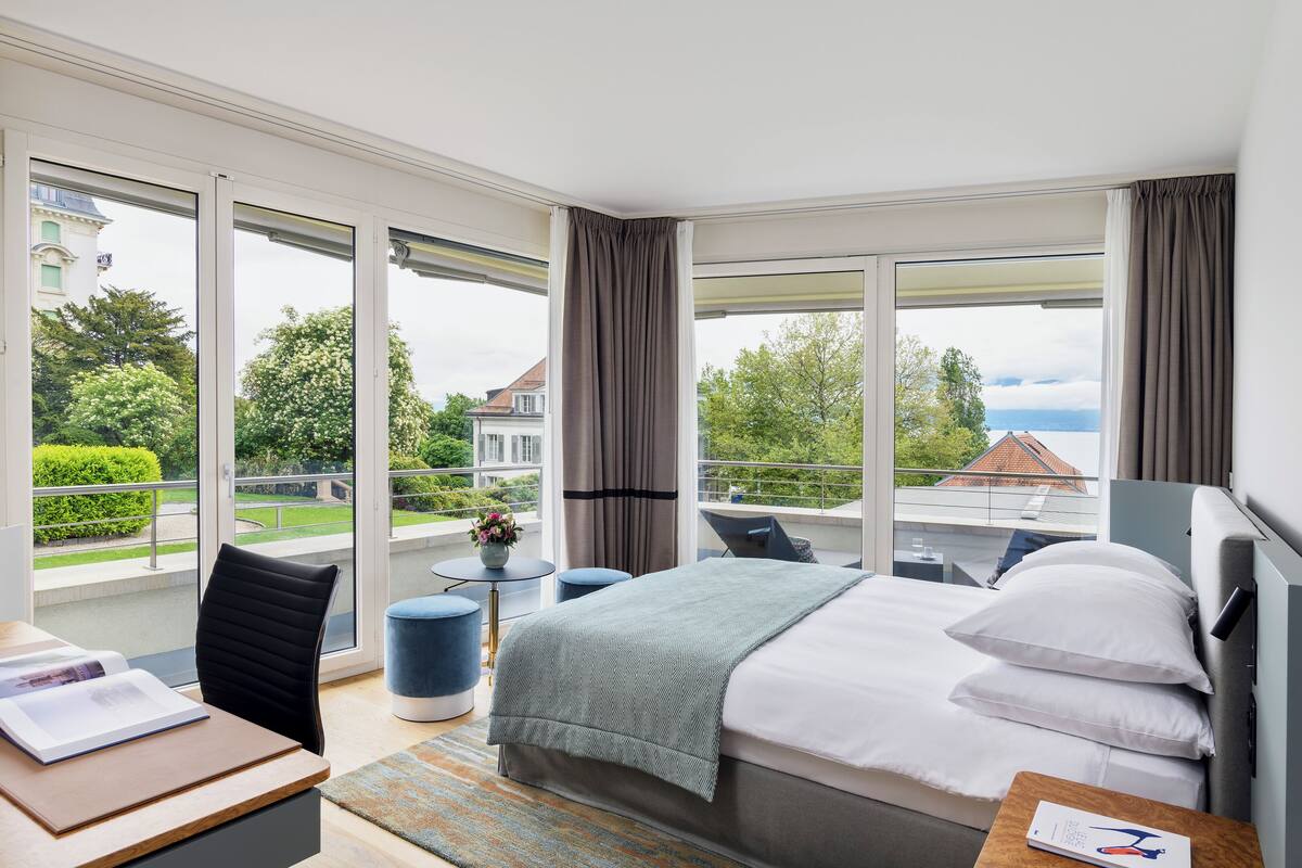 A modern hotel room with a large bed, a desk, and a balcony with a view of greenery and buildings.