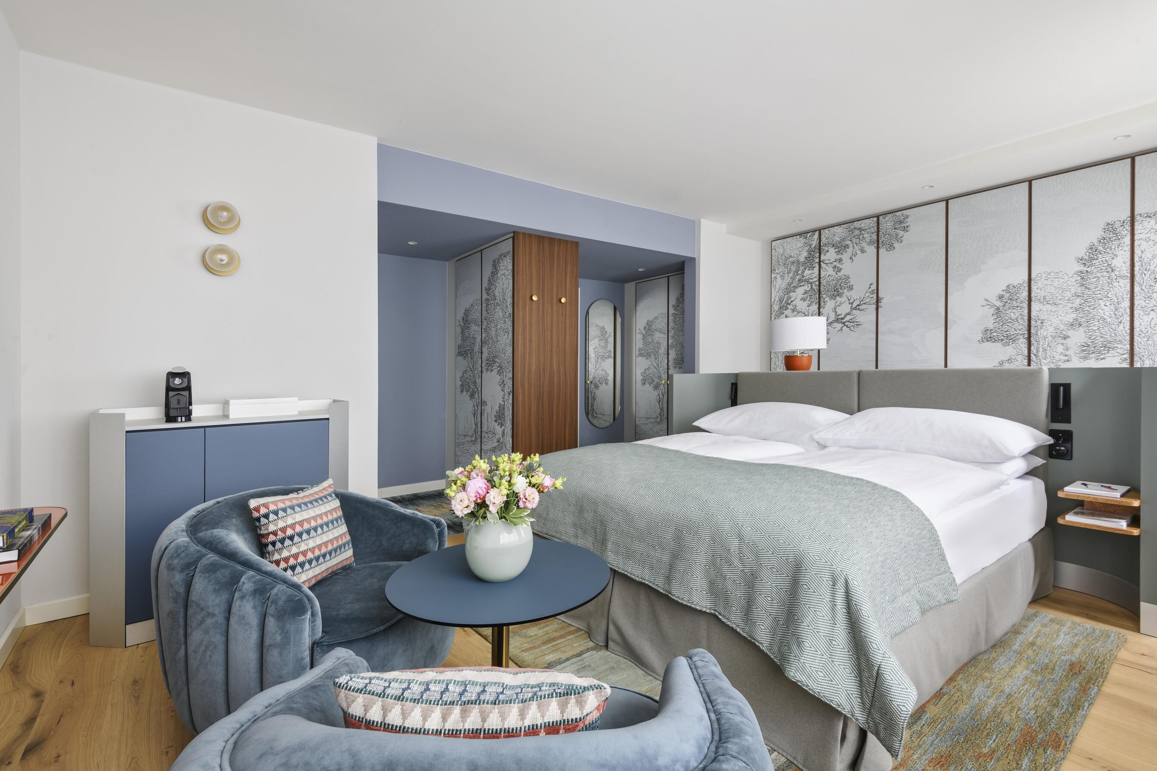 A modern hotel room with a bed, a desk, a chair, a small table with flowers, and a wall with artwork.
