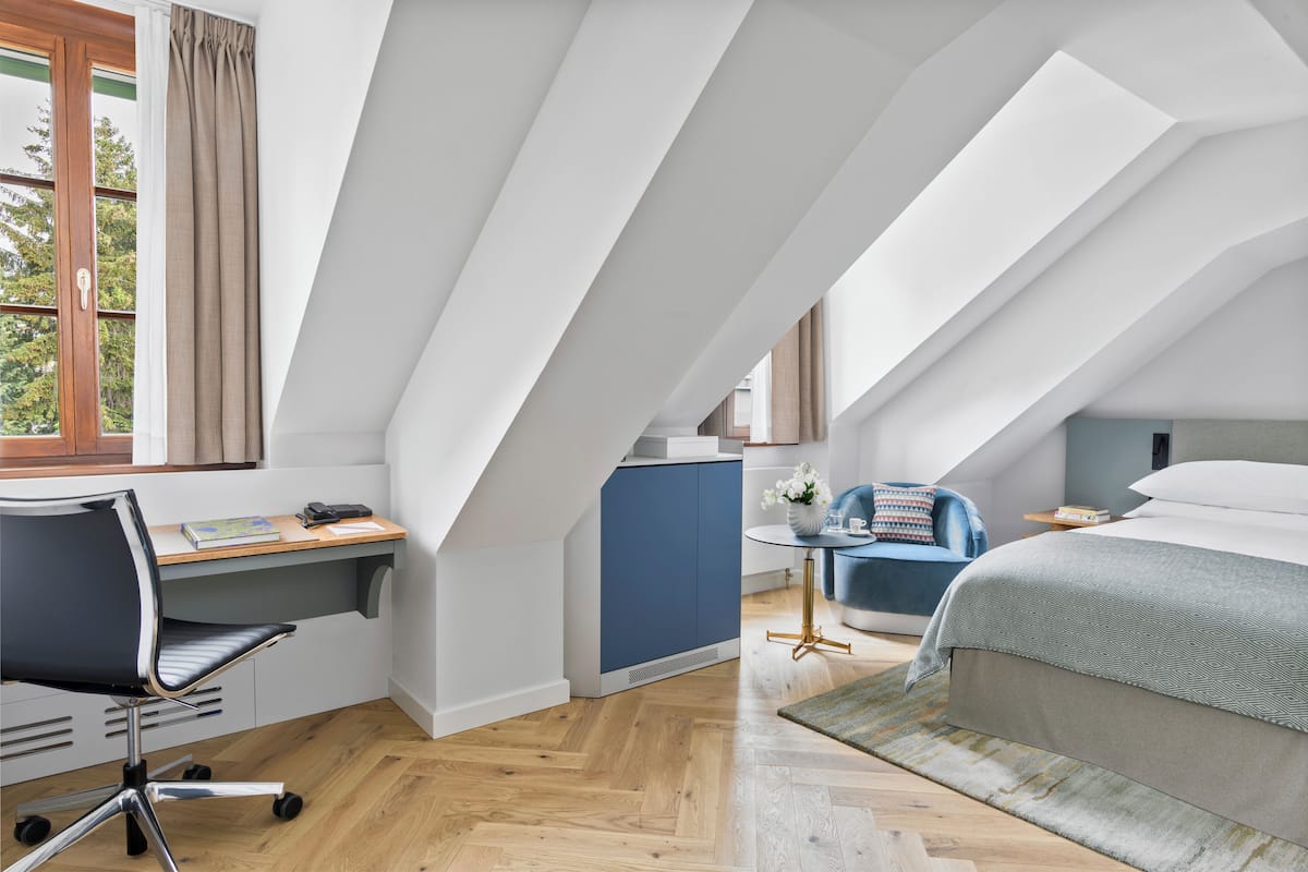 A modern bedroom with a sloped ceiling, a desk, a chair, a bed, and a small table with a vase of flowers.