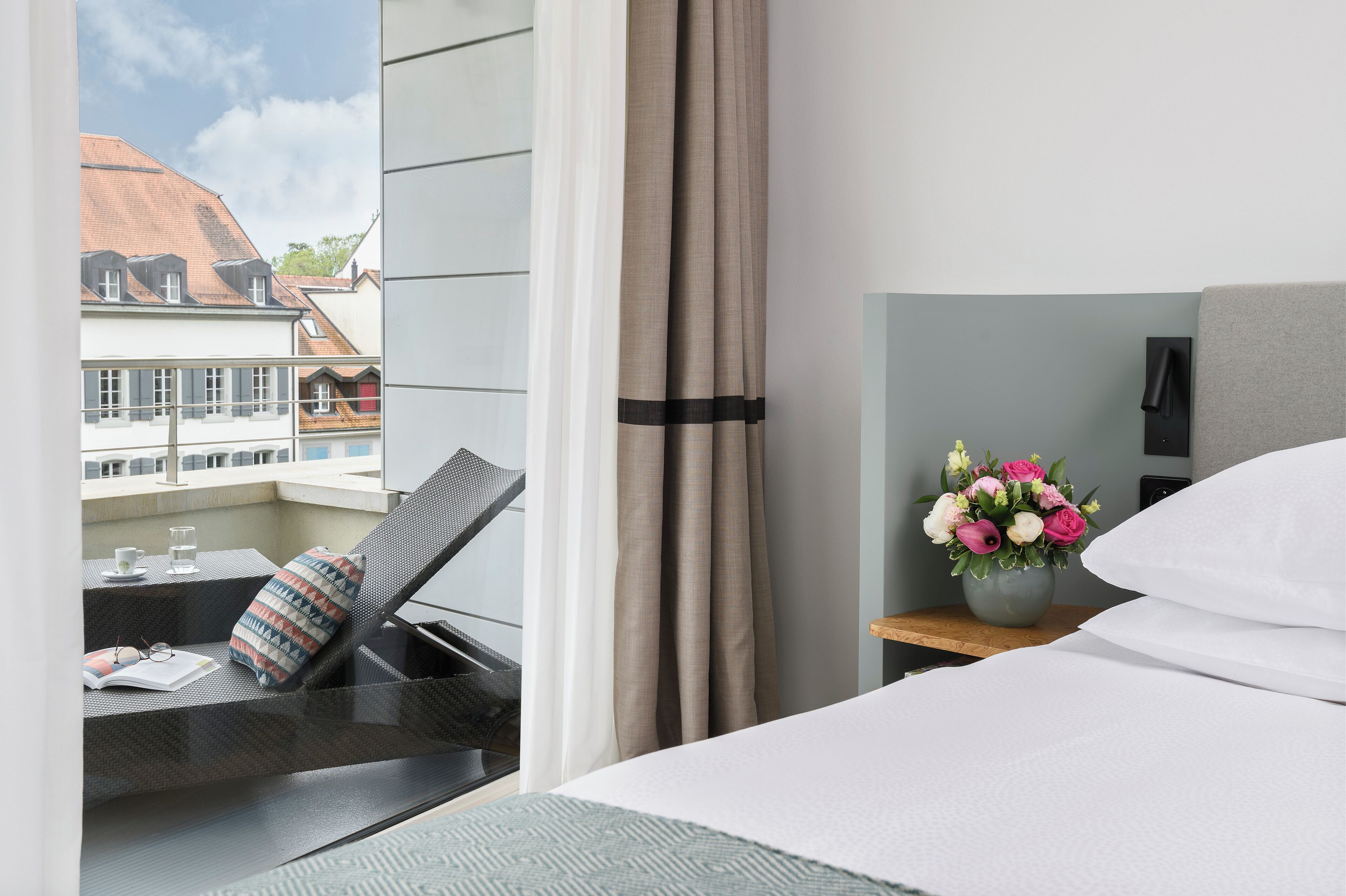 Quarto de hotel com cama, uma pequena mesa com um vaso de flores e vista para edifícios.