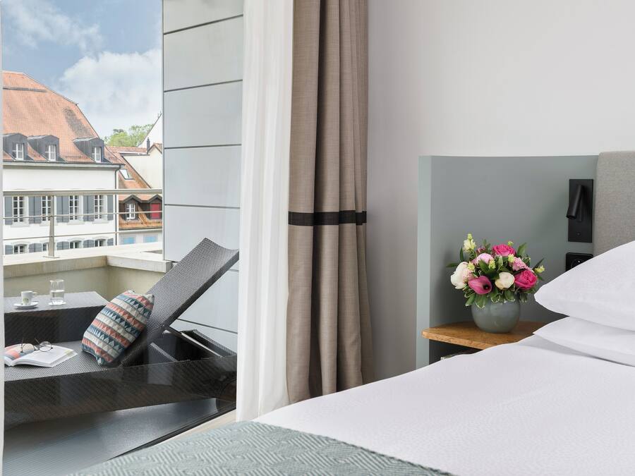 Quarto de hotel com cama, uma pequena mesa com um vaso de flores e vista para edifícios.