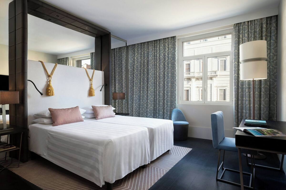 A hotel room with two beds, a desk, a chair, a lamp, and a large window with curtains.