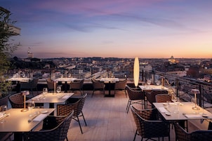 Area pranzo all'aperto con tavoli e sedie, affacciata su una città al tramonto.