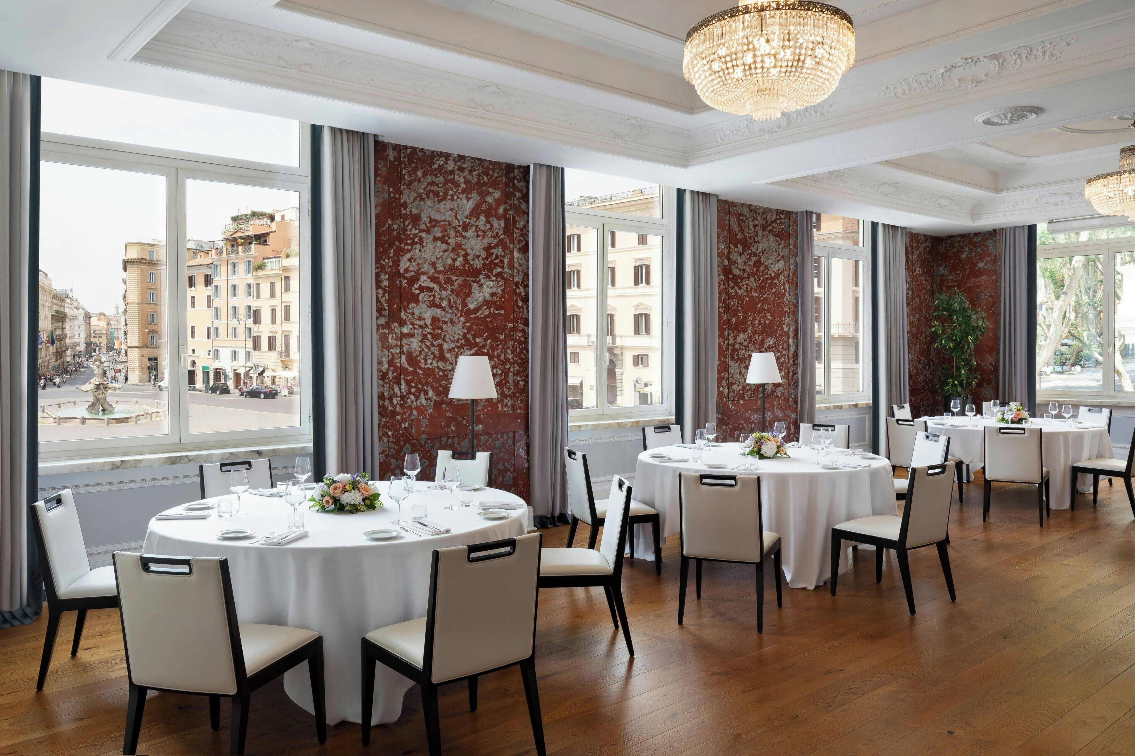 A restaurant with round tables set for dining, large windows with city views, and elegant chandeliers.